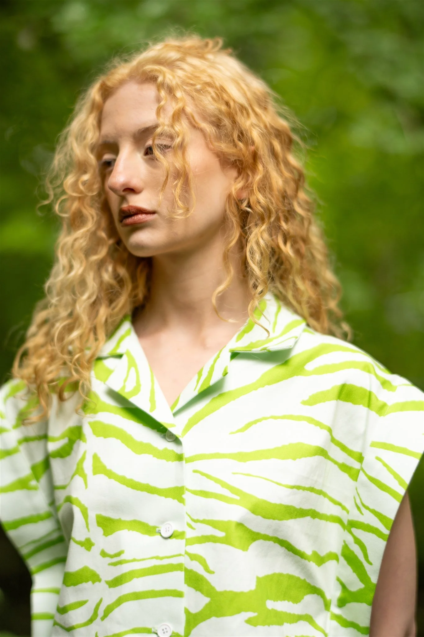 A woman with curly blonde hair wearing a short-sleeve button-up shirt with green and white zebra stripes outdoors with green foliage in the background.