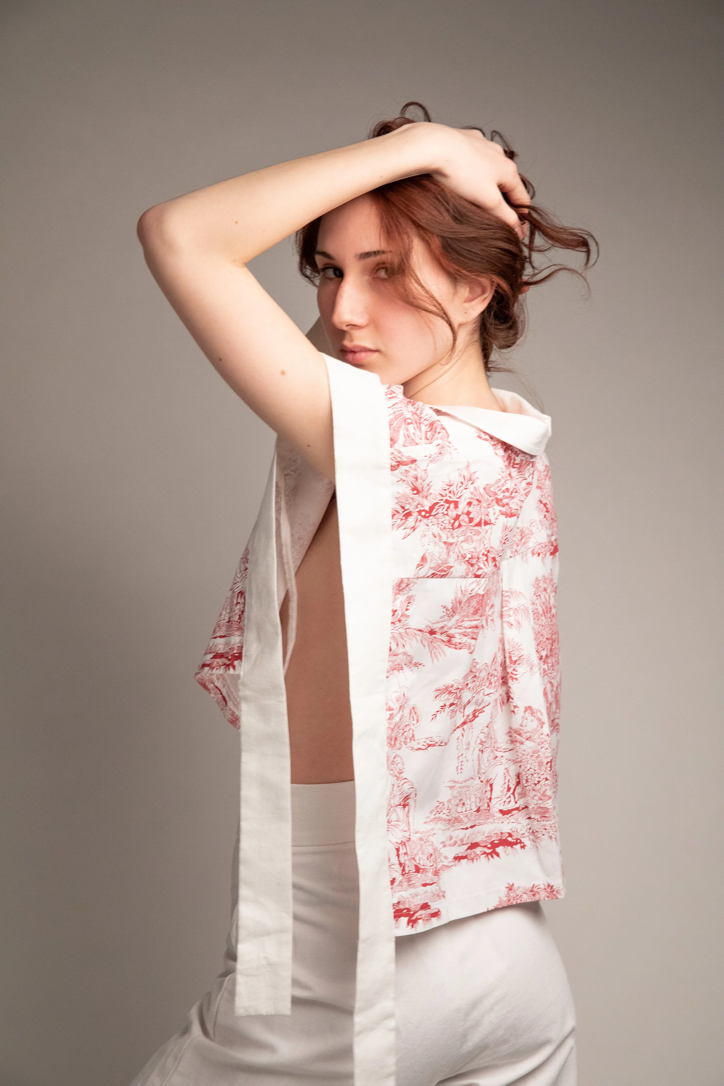 A young woman with short red hair is posing, with her left arm raised and her hand in her hair, wearing a sleeveless patterned shirt and white pants, against a neutral background.