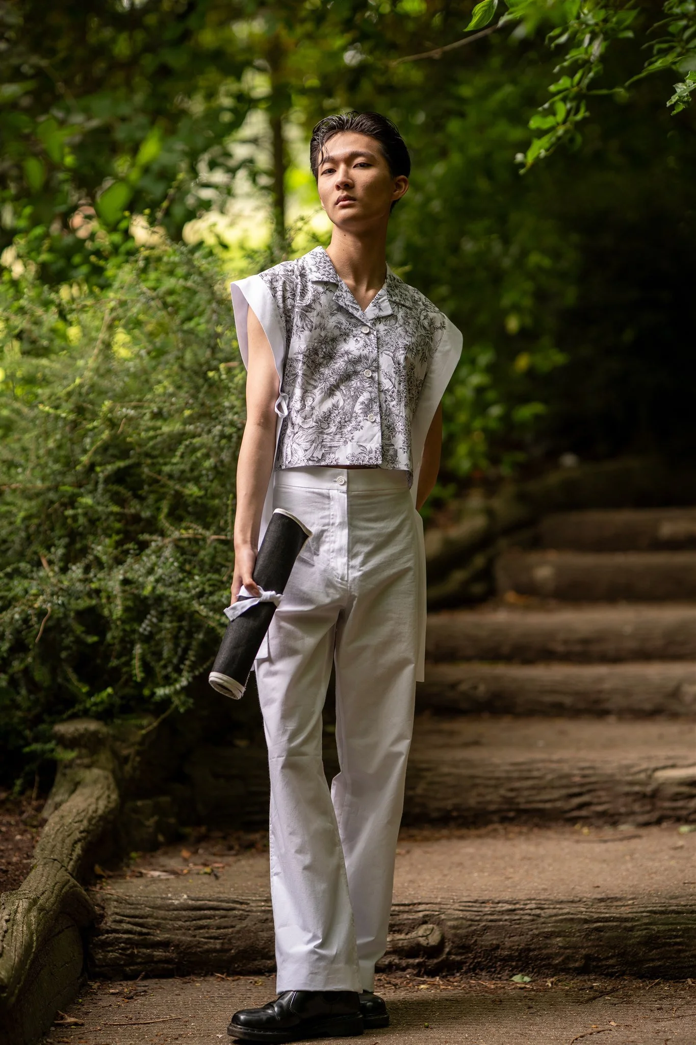 A person standing outdoors on a trail surrounded by greenery, wearing white pants, black shoes, and a sleeveless patterned top, holding a rolled-up mat.