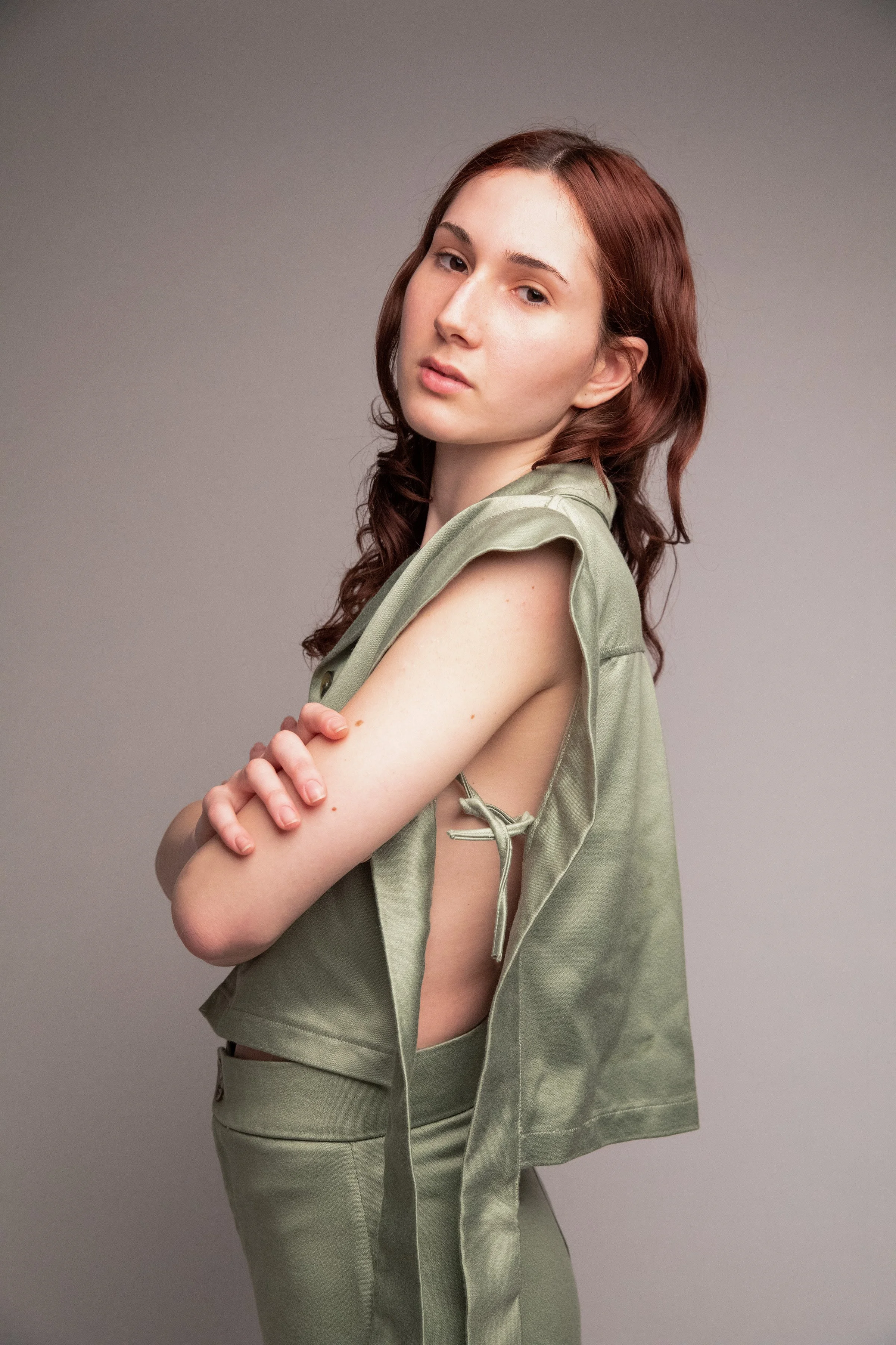 A young woman with red hair and fair skin is standing against a neutral gray background. She is wearing a sleeveless, olive-green top with side ties and matching pants. Her arms are crossed, and she gazes directly at the camera with a neutral express