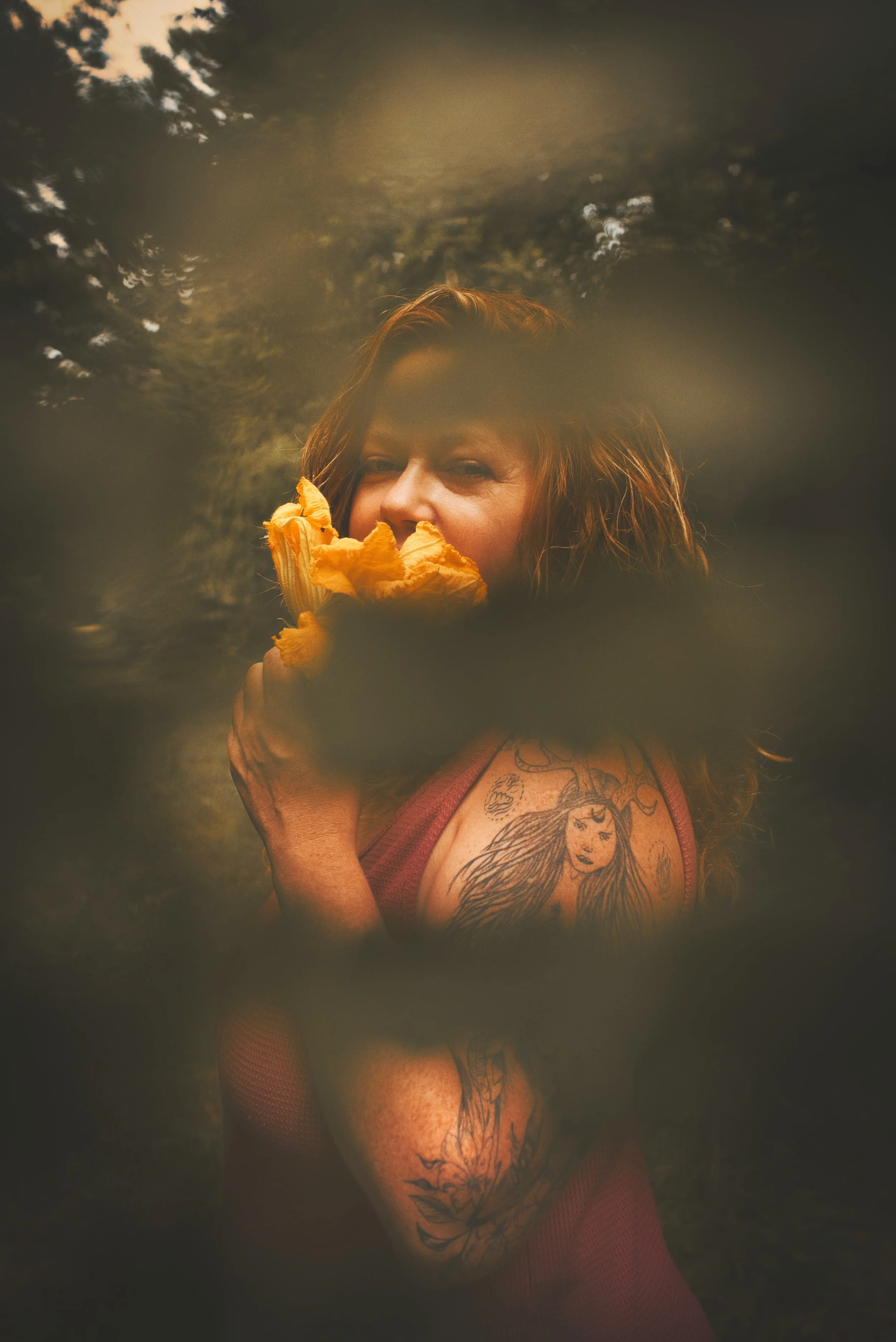 Portrait d'une femme rousse dans les bois tenant une fleur de courgette, et avec une feuille de fougère devant l'objectif