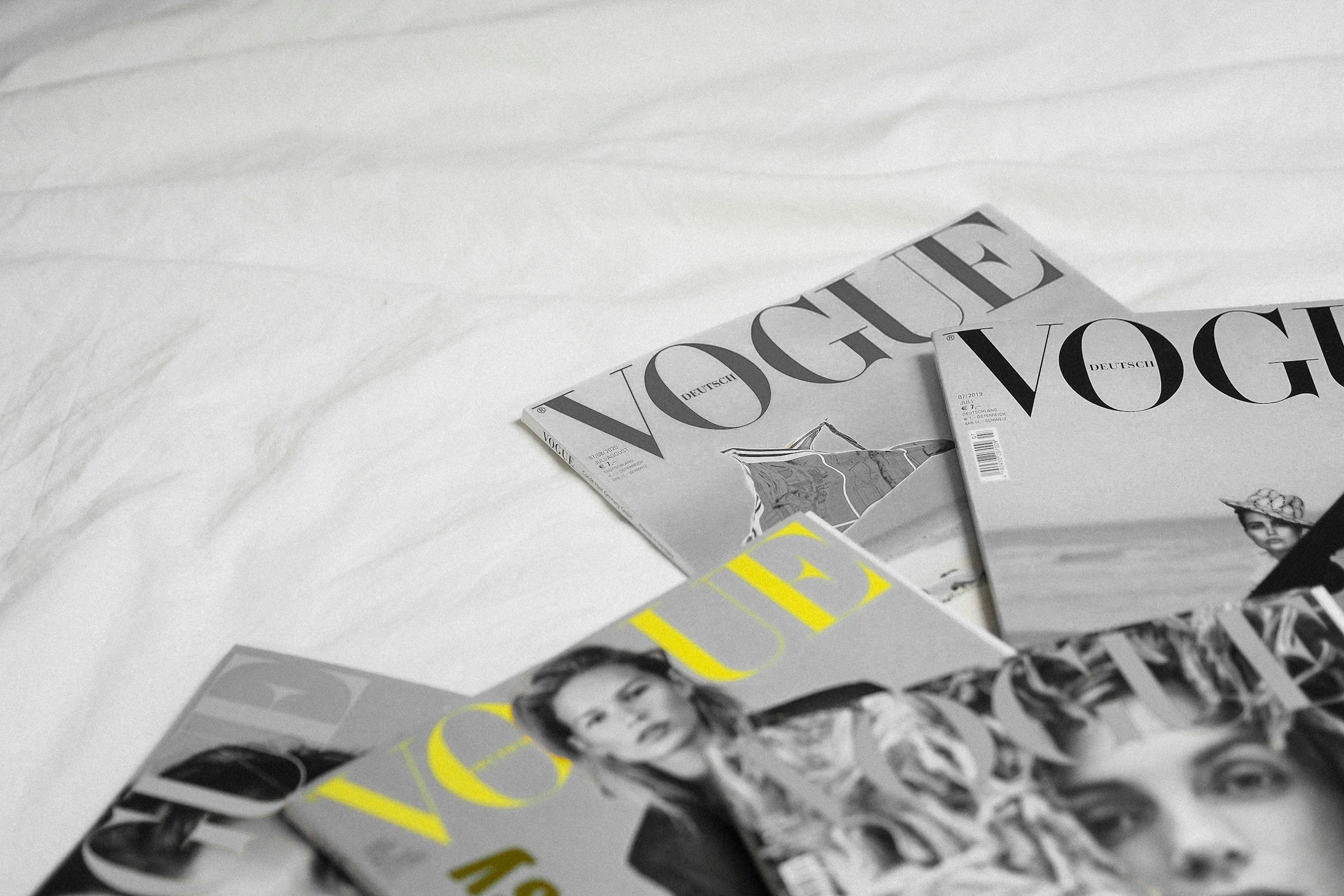 Magazines Vogue et Vogue Deutschland empilés sur un lit blanc.