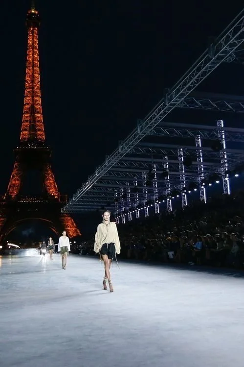 Défilé de mode en plein air avec la Tour Eiffel illuminée en arrière-plan, modèles marchant sur le runway nocturne, public assis sur les côtés.