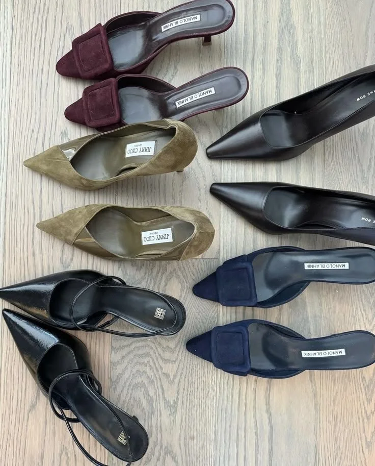 Six paires de chaussures à talons alignées sur un sol en bois, incluant des escarpins en daim bordeaux, en cuir beige, en cuir noir verni, et en velours bleu.
