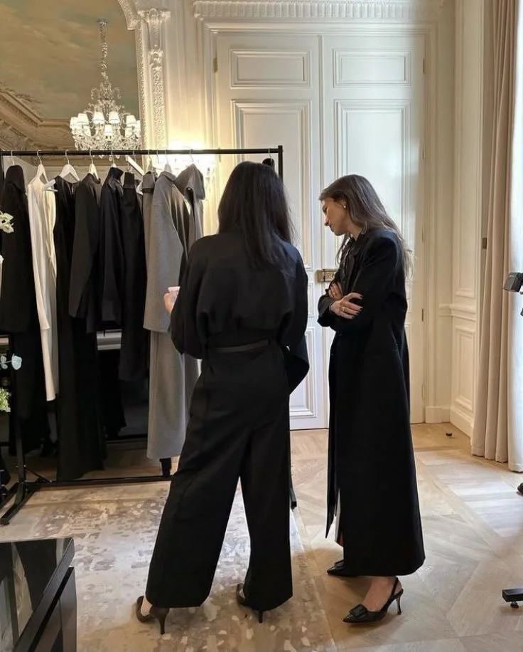 Deux femmes discutant dans une pièce élégante avec un portant de vêtements sombres, une grande porte blanche, et un lustre en cristal suspendu au plafond.