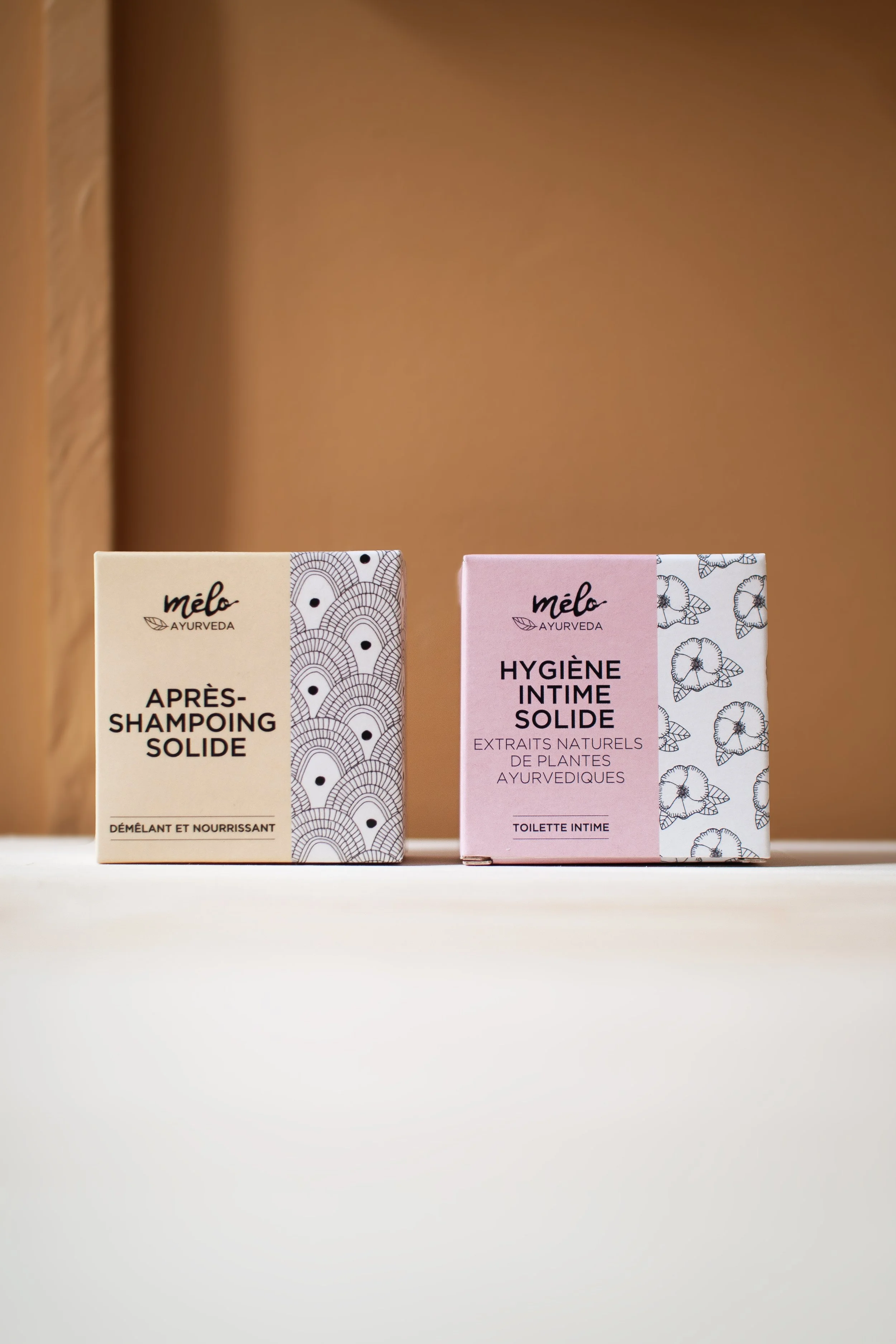 Deux boîtes de produits de soins intimes, l'une pour après-shampoing solide et l'autre pour hygiène intime solide, posées sur une surface blanche avec un fond beige.