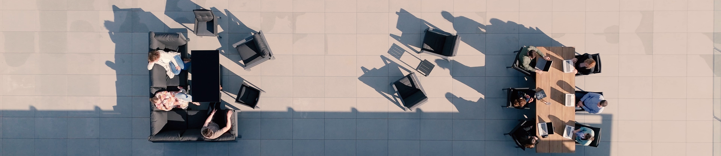Top-down view of six people working on laptops and engaging in a meeting outdoors on a tiled surface, with multiple chairs and a sofa arranged around a table.