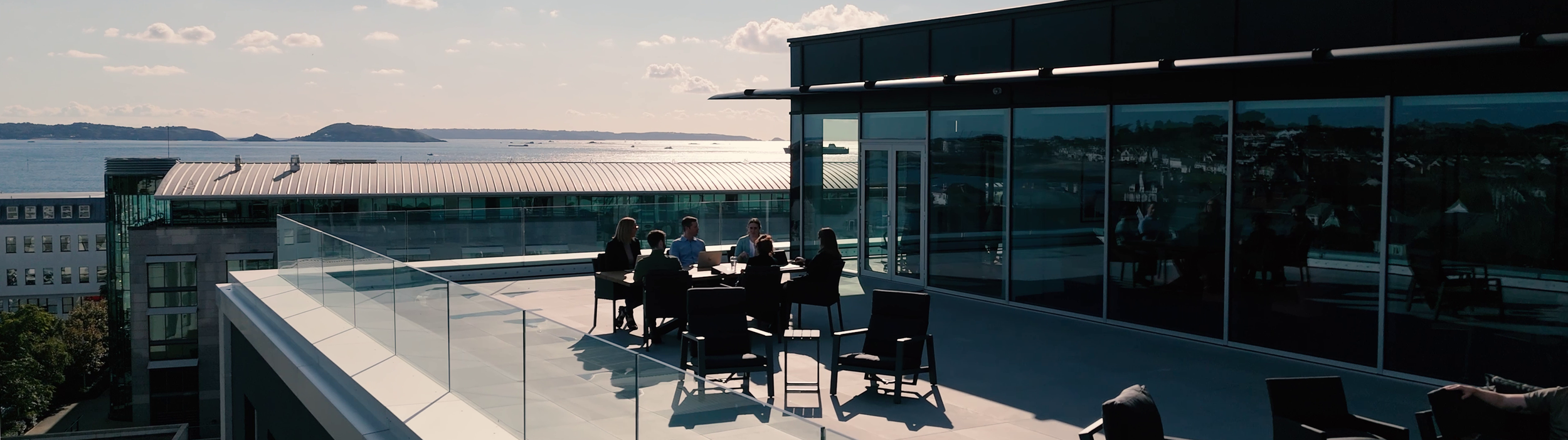 Business meeting on a rooftop terrace overlooking a bay, with six people seated at a table with laptops, glass railings, and a view of the water and islands in the background.