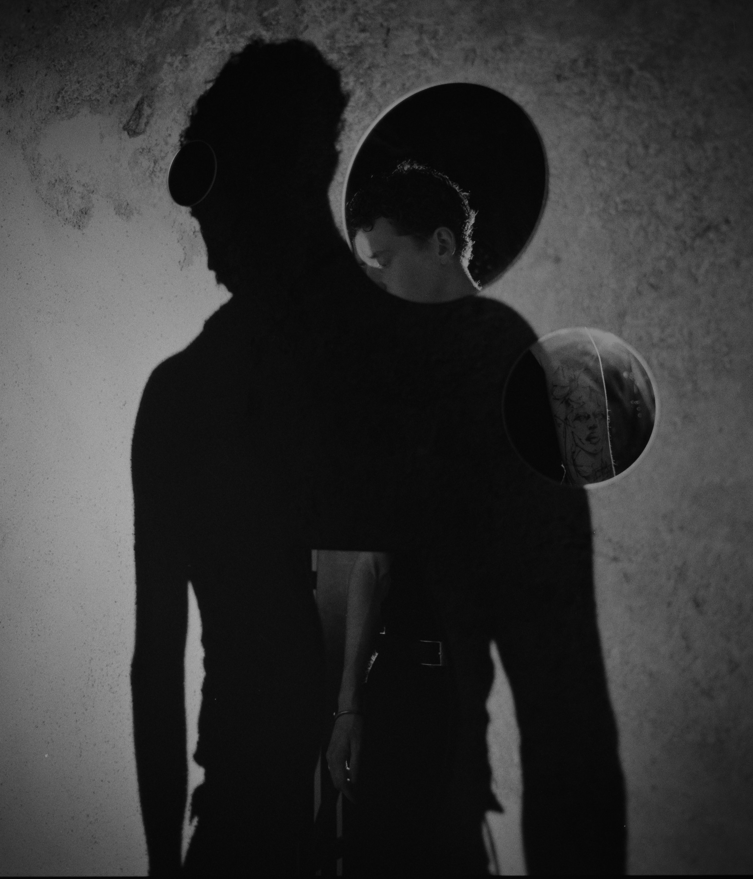 Schwarz-Weiß-Foto eines Menschen, der seinen Schatten an einer Wand wirft, wobei der Schatten ein Gesicht auf einer Linie sichtbar macht, und eine Person, die in einem Spiegel einen Teil ihres Gesichts sieht.