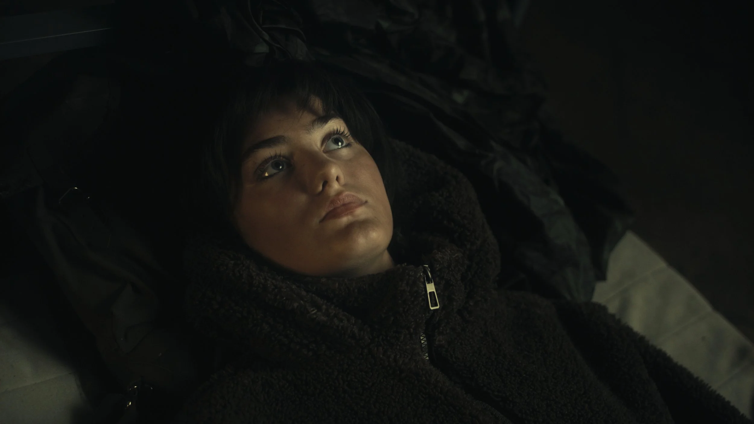 Nahaufnahme eines jungen Mannes mit dunklen Haaren, der auf dem Rücken liegend auf einem dunklen Untergrund schaut.