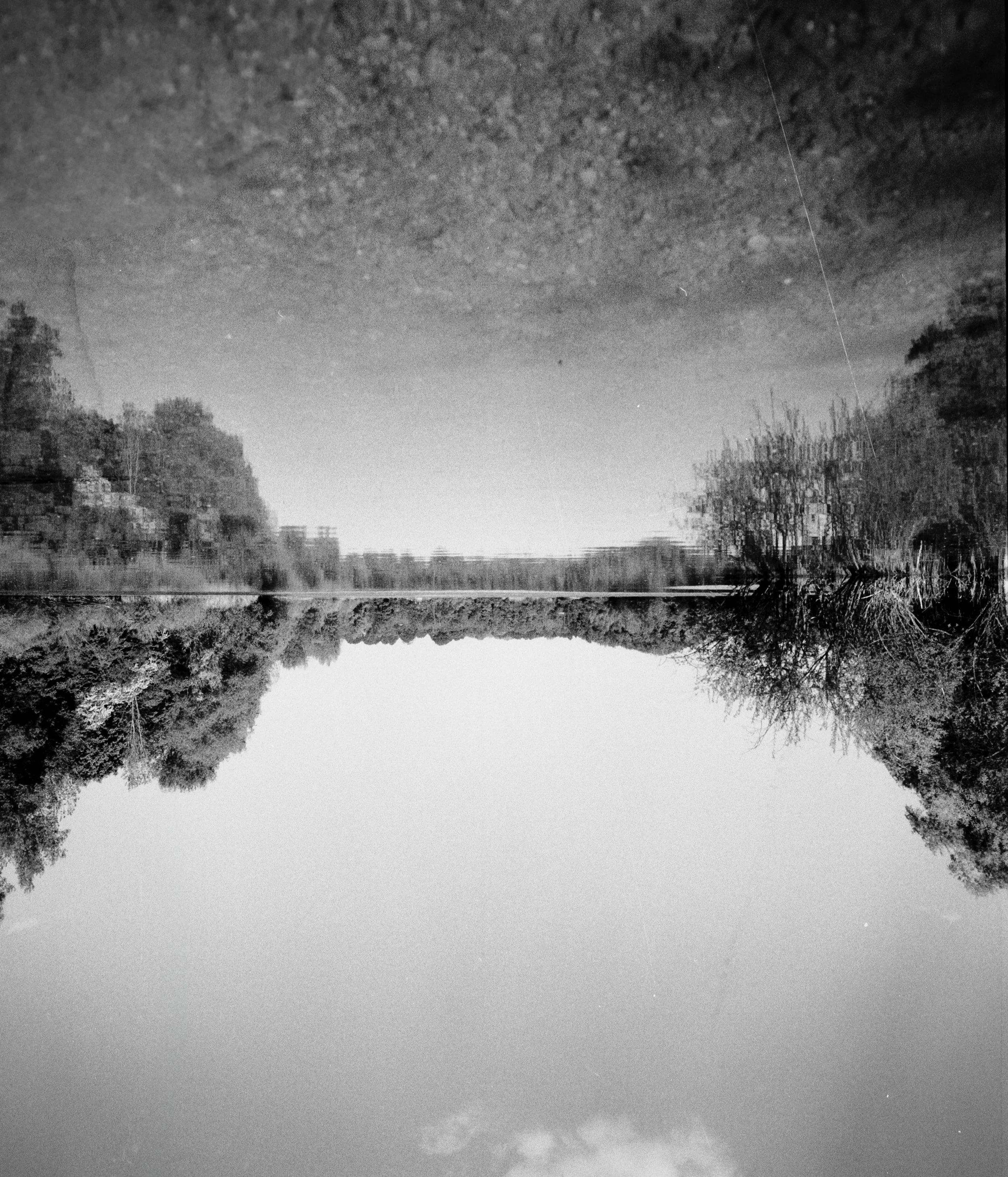 Schwarz-Weiß-Foto eines ruhigen Flusses, umgeben von Bäumen, mit einem bewölkten Himmel darüber.