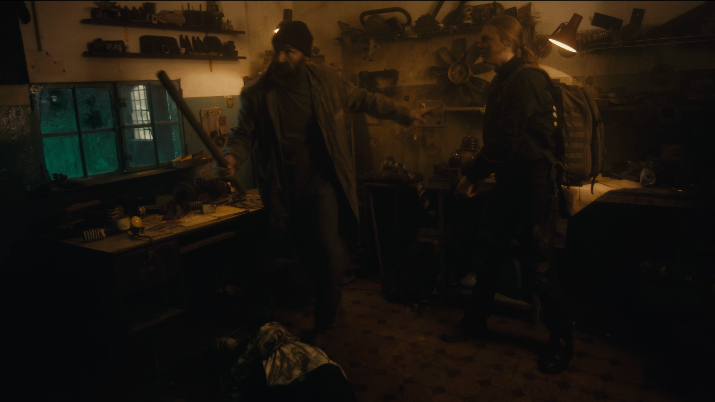 Zwei Personen in einer Werkstatt, eine hält einen Baseballschläger, die andere trägt einen Rucksack.