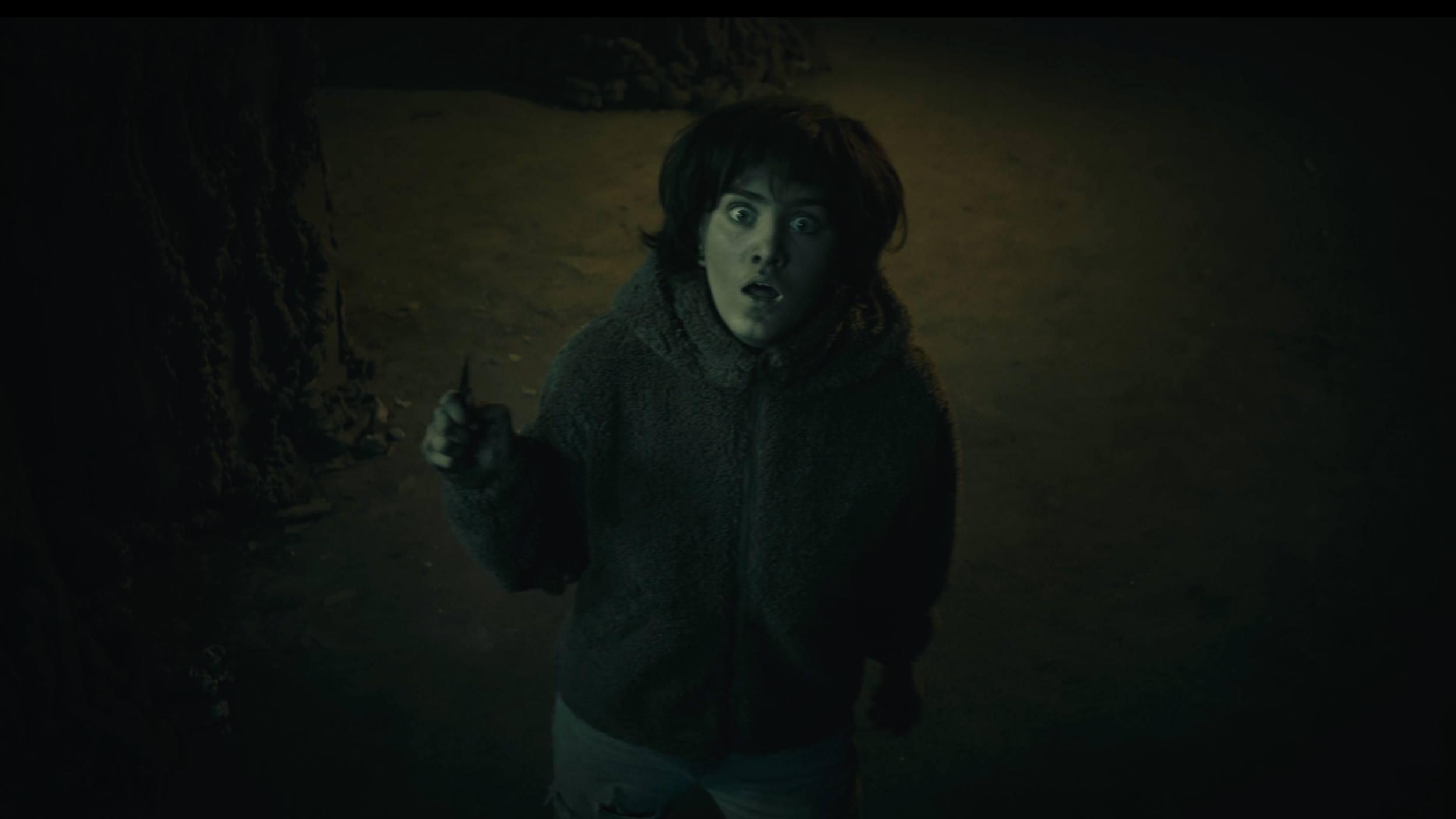 Junge Person mit dunklen Haaren trägt eine dunkle Jacke und steht im Schatten, der Blick ist erstaunt oder schockiert, die Umgebung ist dunkel und unheimlich.