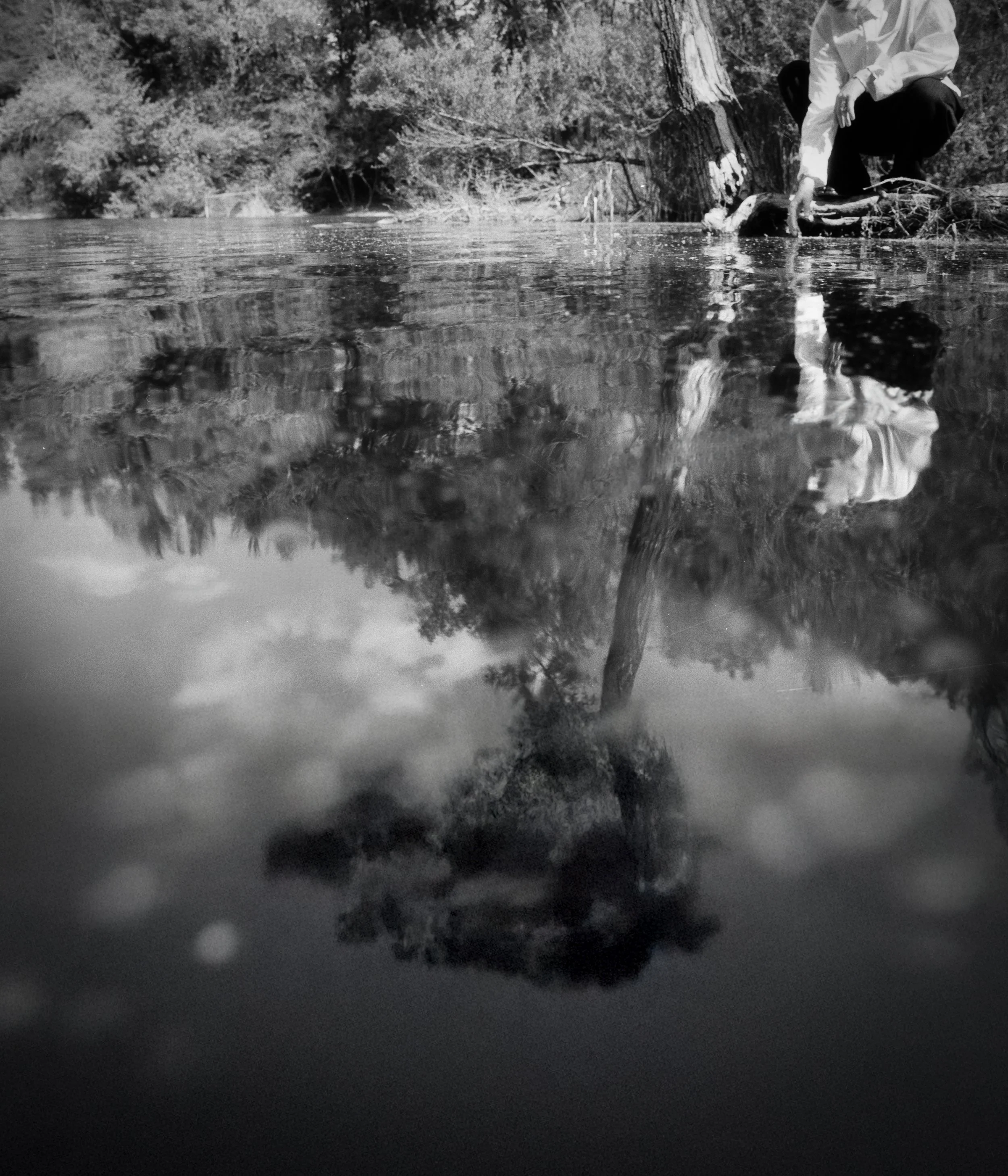 Schwarz-Weiß-Foto eines Menschen, der an einem Fluss oder See steht und den Wasserstand berührt, mit Baumstämmen und Bäumen im Hintergrund und deren Reflexion im Wasser.