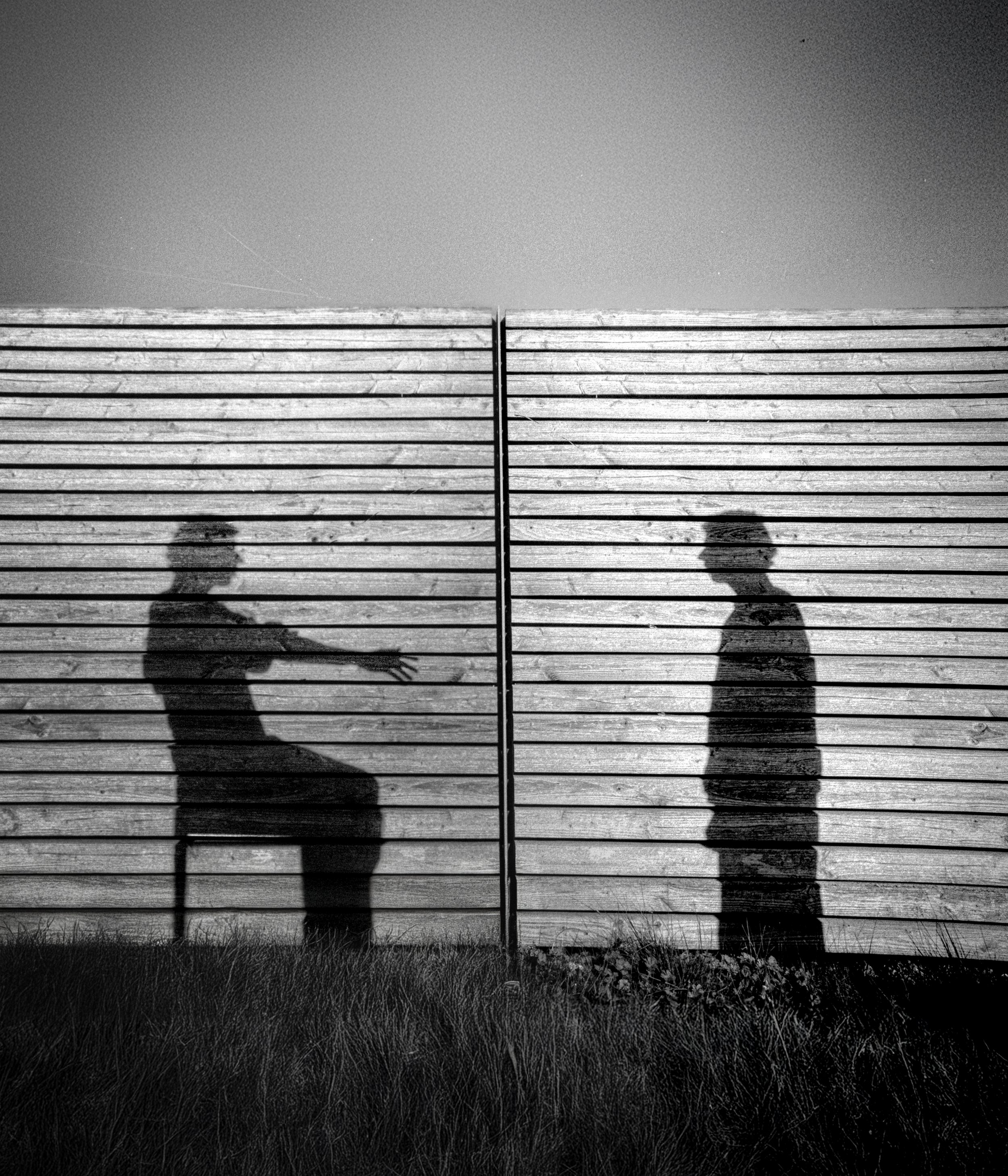 Schattenspiel von zwei Personen hinter einem Zaun mit horizontalen Holzlatten, eine Person sitzt auf einem Stuhl und reicht die Hand durch die Latten, die andere Person steht, alle in Schwarz-Weiß.