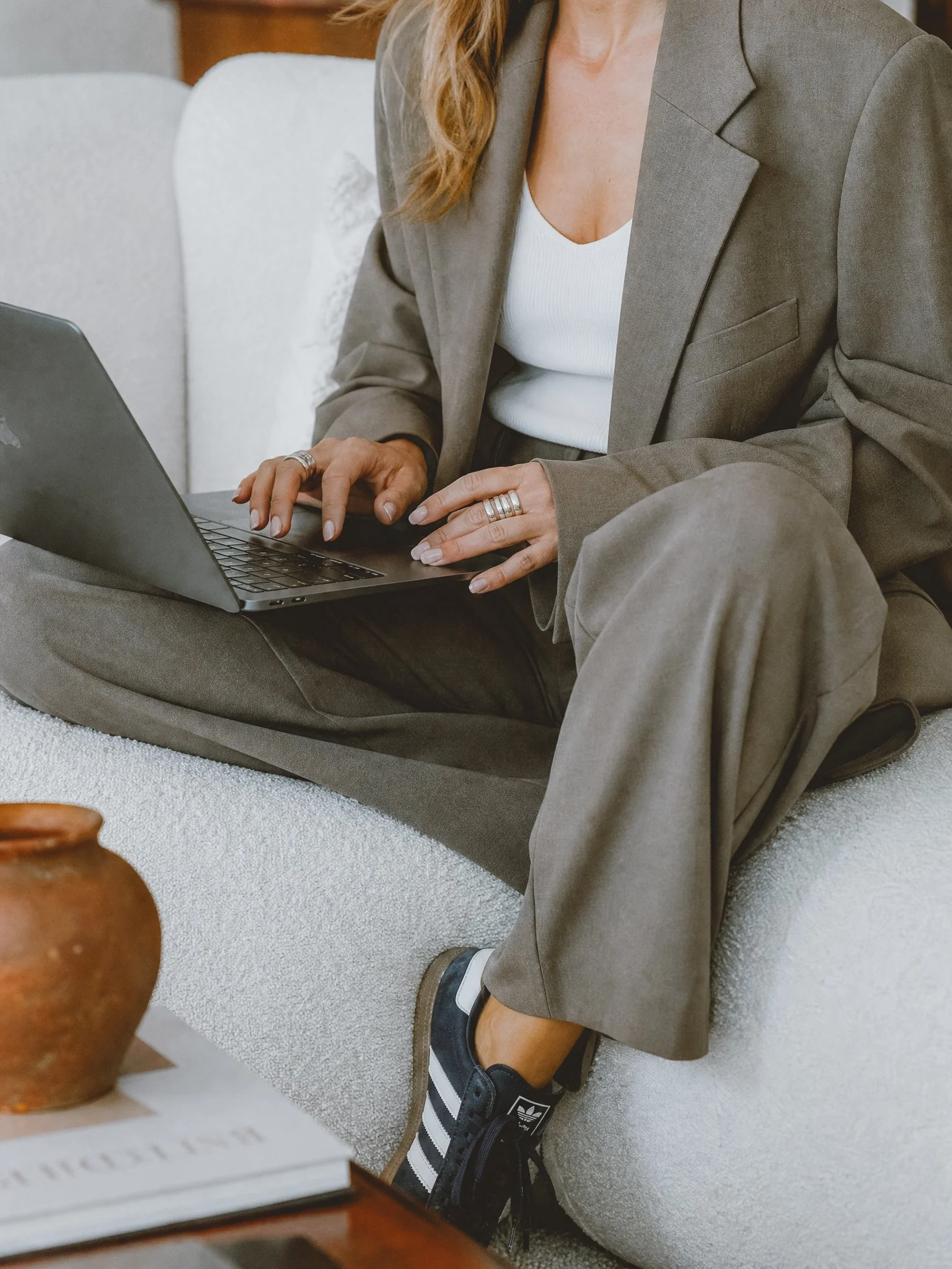 Person wearing a gray suit jacket and white shirt, sitting cross-legged on a soft surface, typing on a laptop with a wooden table nearby.