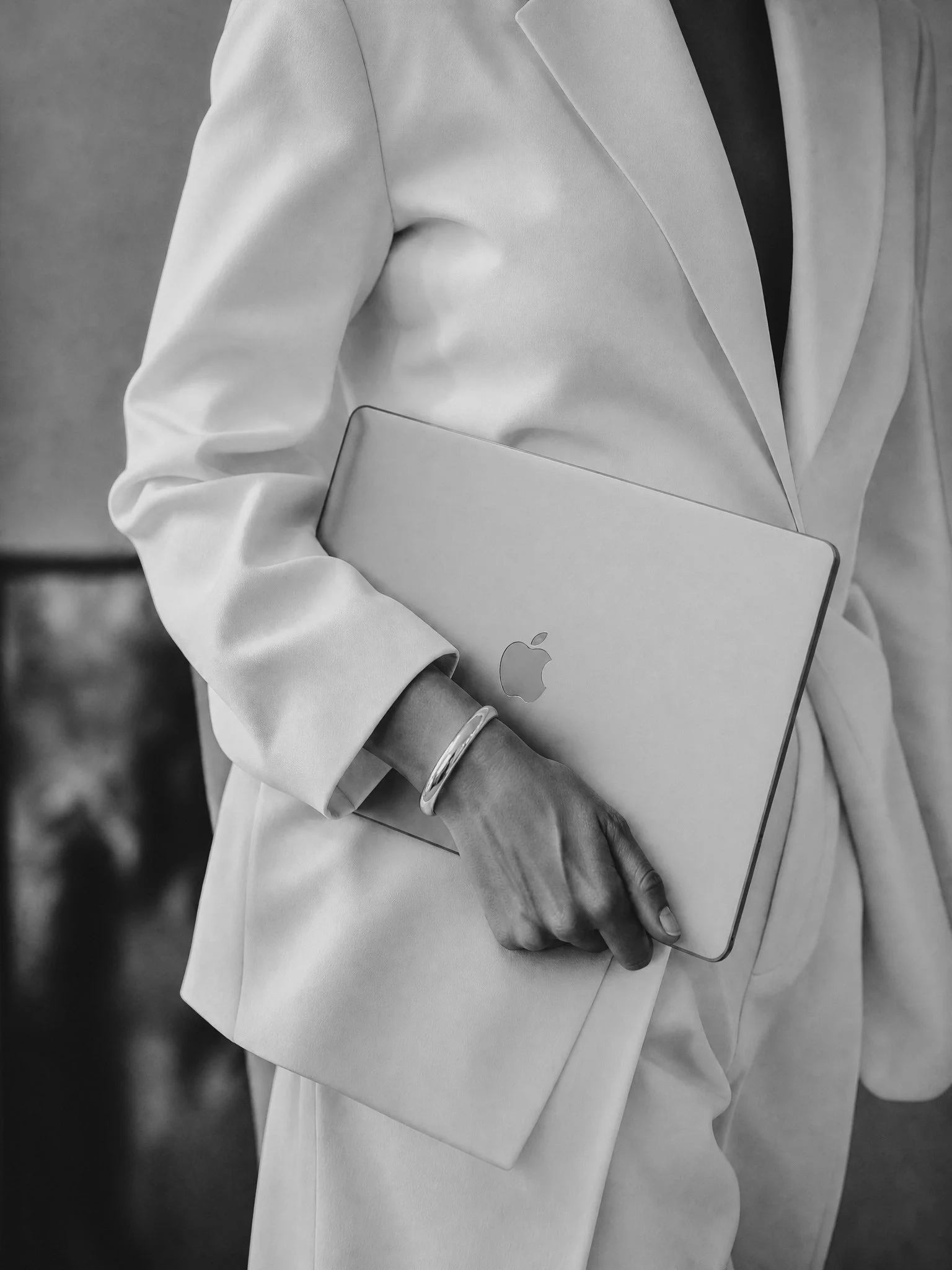 Close-up of a person dressed in a white blazer holding a closed silver MacBook laptop.