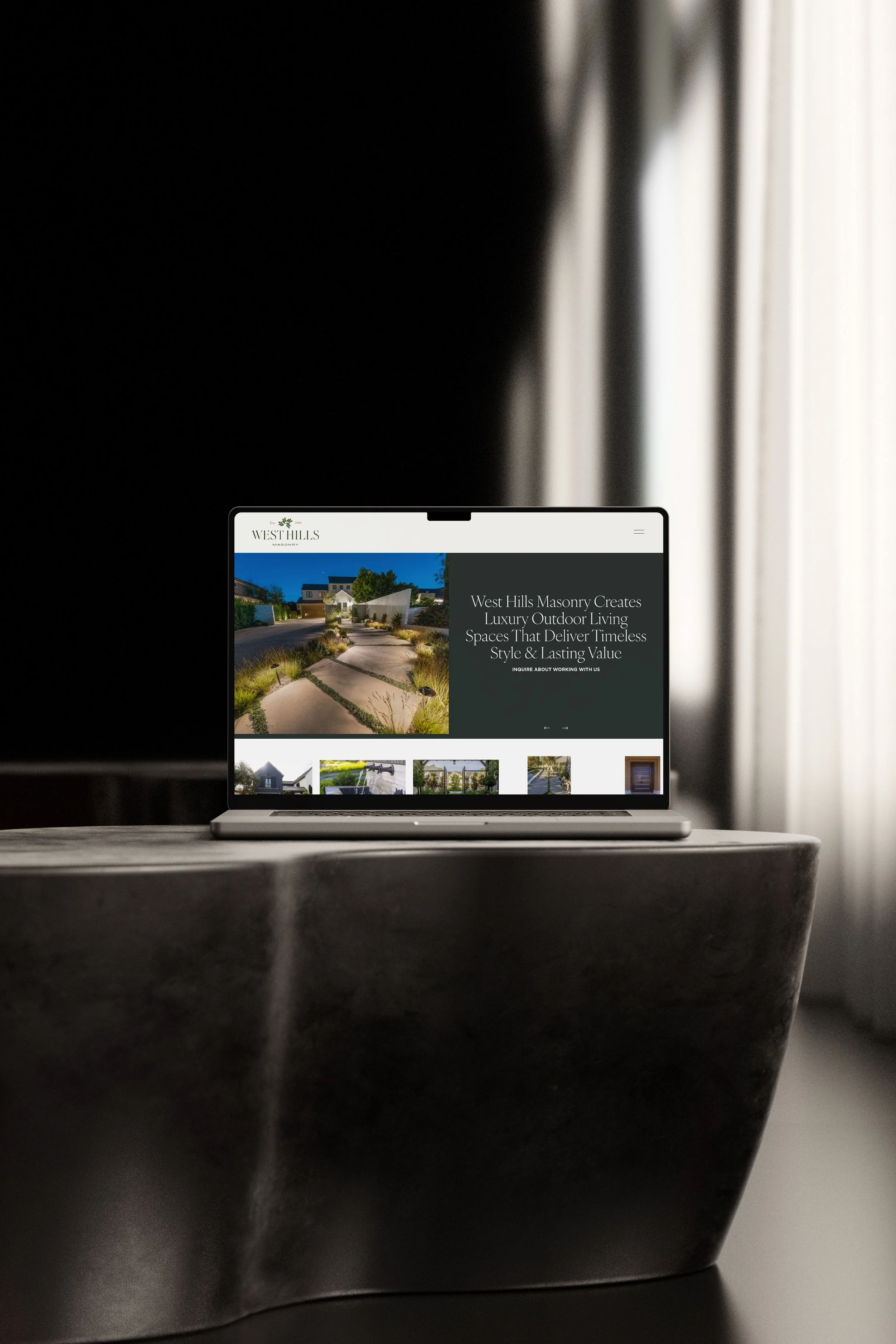 Laptop on a dark table displaying a website for West Hills Masonry, featuring images of outdoor landscaping and masonry projects.