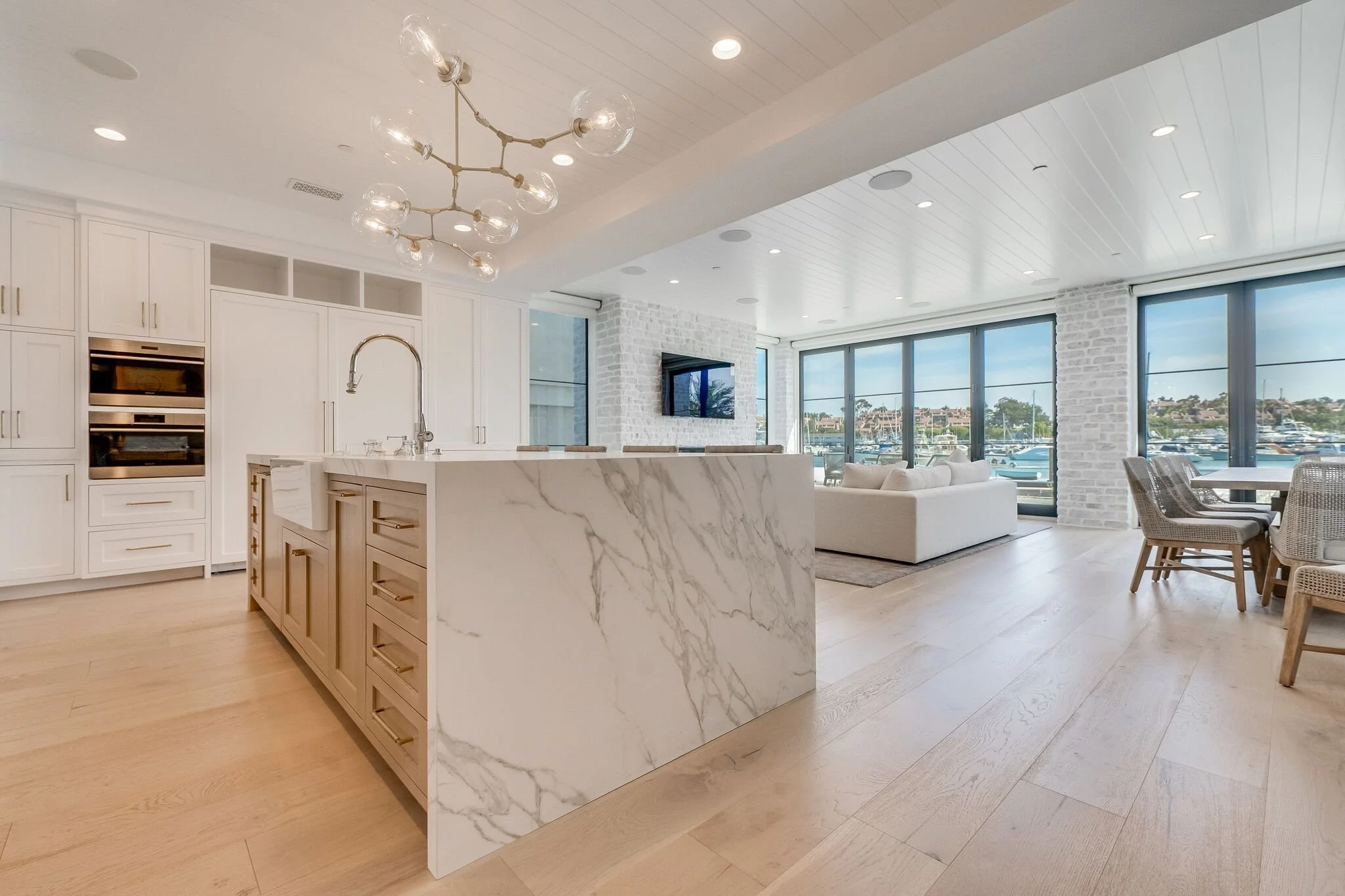 Modern open-concept kitchen and living room with white cabinetry, marble island, light wood floors, large windows overlooking a marina, and a white brick accent wall.