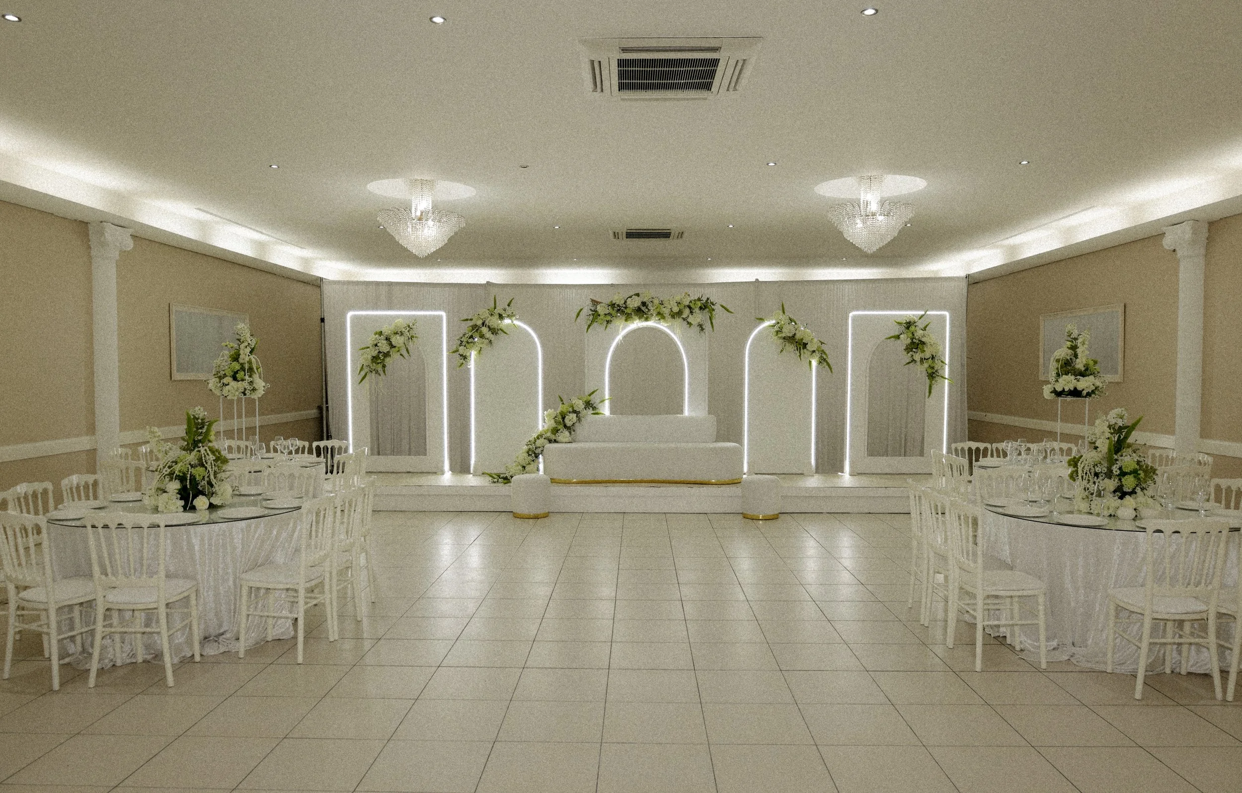 Een elegante witte feestzaal met round tafels, versierd met bloemstukken, en een prachtige witte achtergrond met verlichte bogen en bloemen voor een bruiloft.
