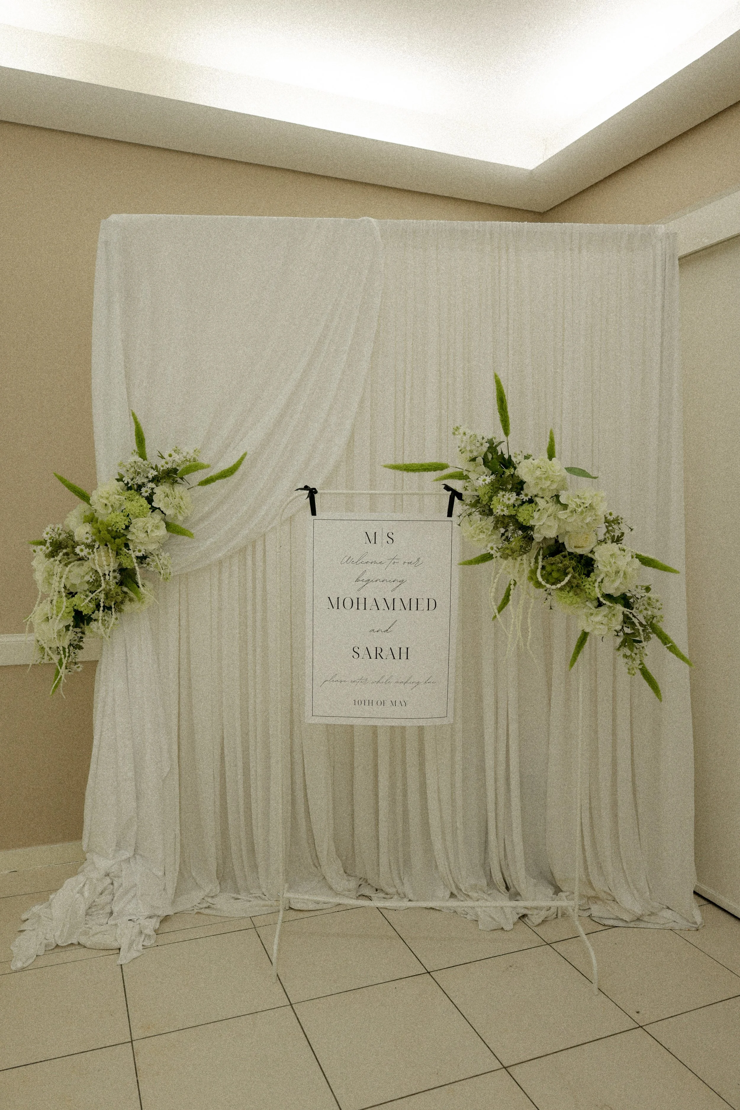 Een witte decoratieve achtergrond met bloemen en een bord met namen en tekst voor een bruiloft.