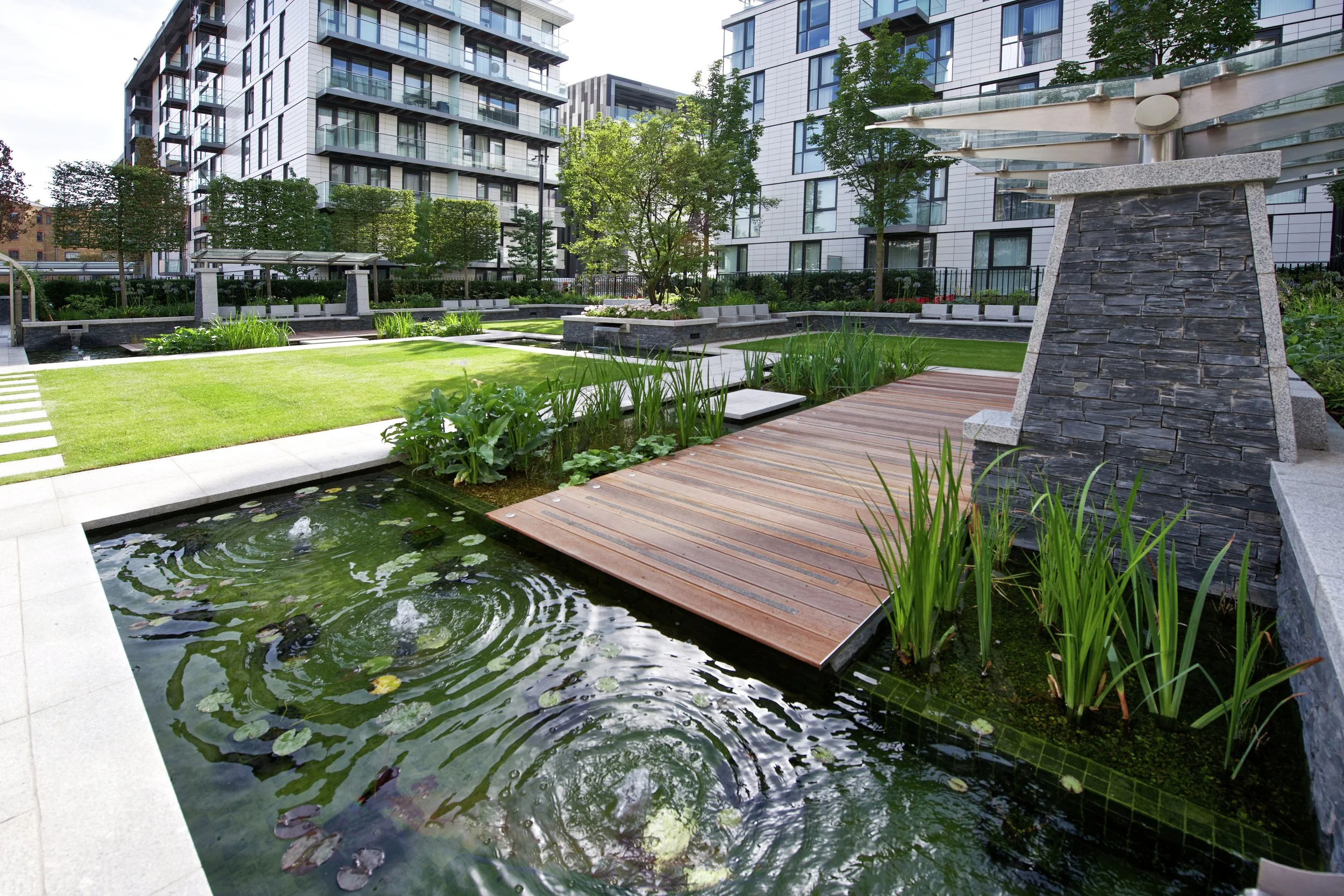 Modern urban green space with a small pond, wooden deck, and lush trees surrounded by tall apartment buildings.
