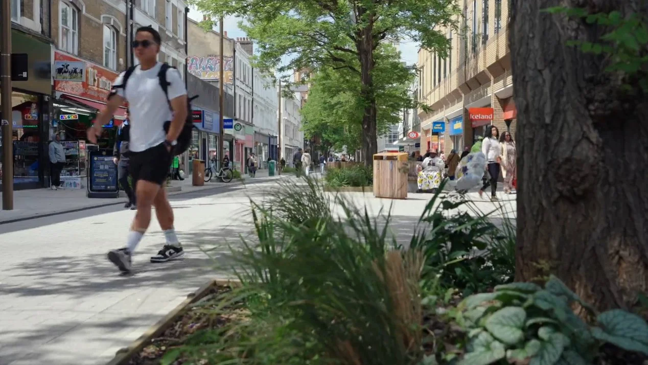 Street scene with pedestrians walking along a sidewalk lined with trees and storefronts, some people sitting outside, and a person in the foreground wearing a white t-shirt and black shorts carrying a backpack.