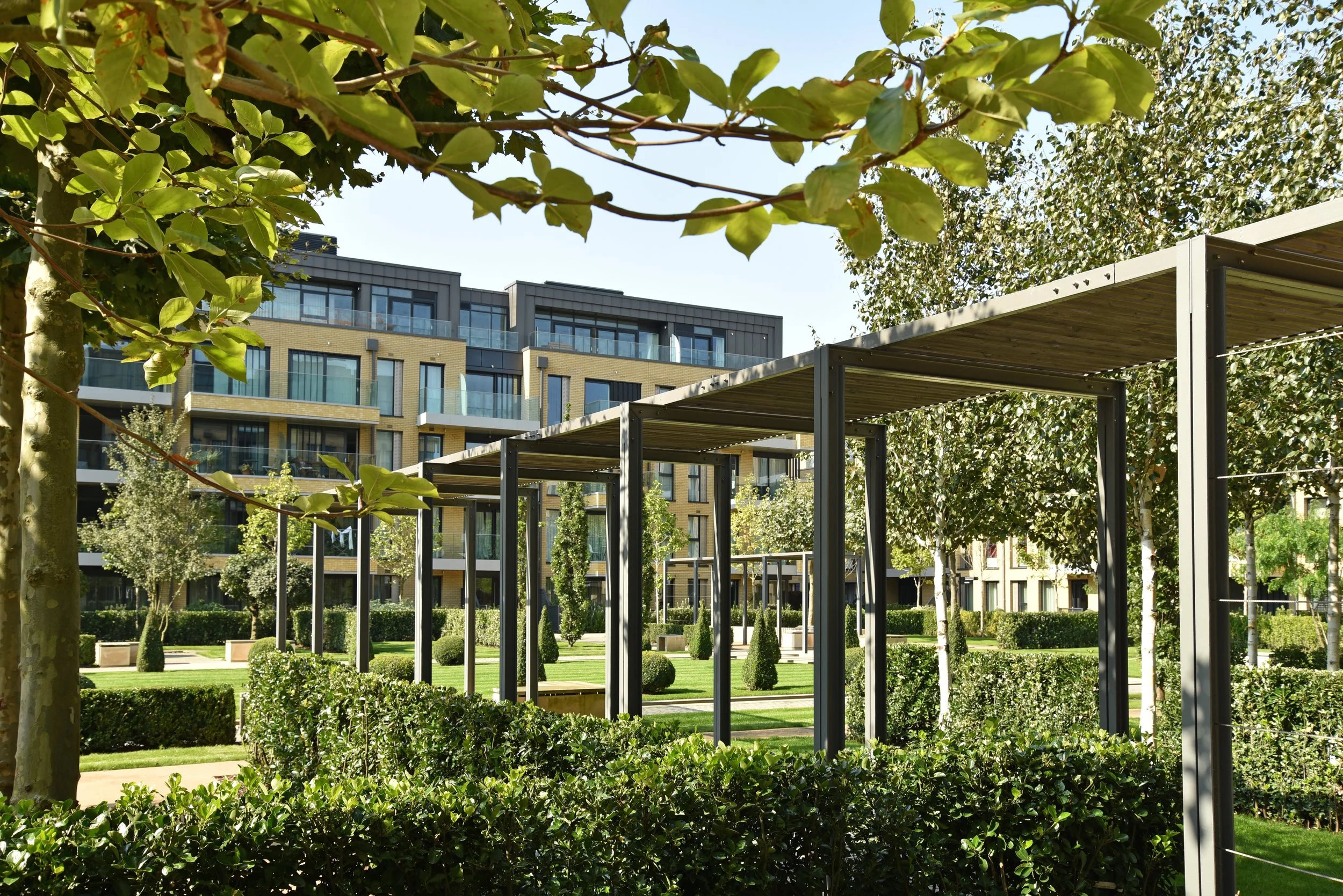 A landscaped urban park with modern apartment buildings in the background, featuring trees, bushes, and metal pergolas.