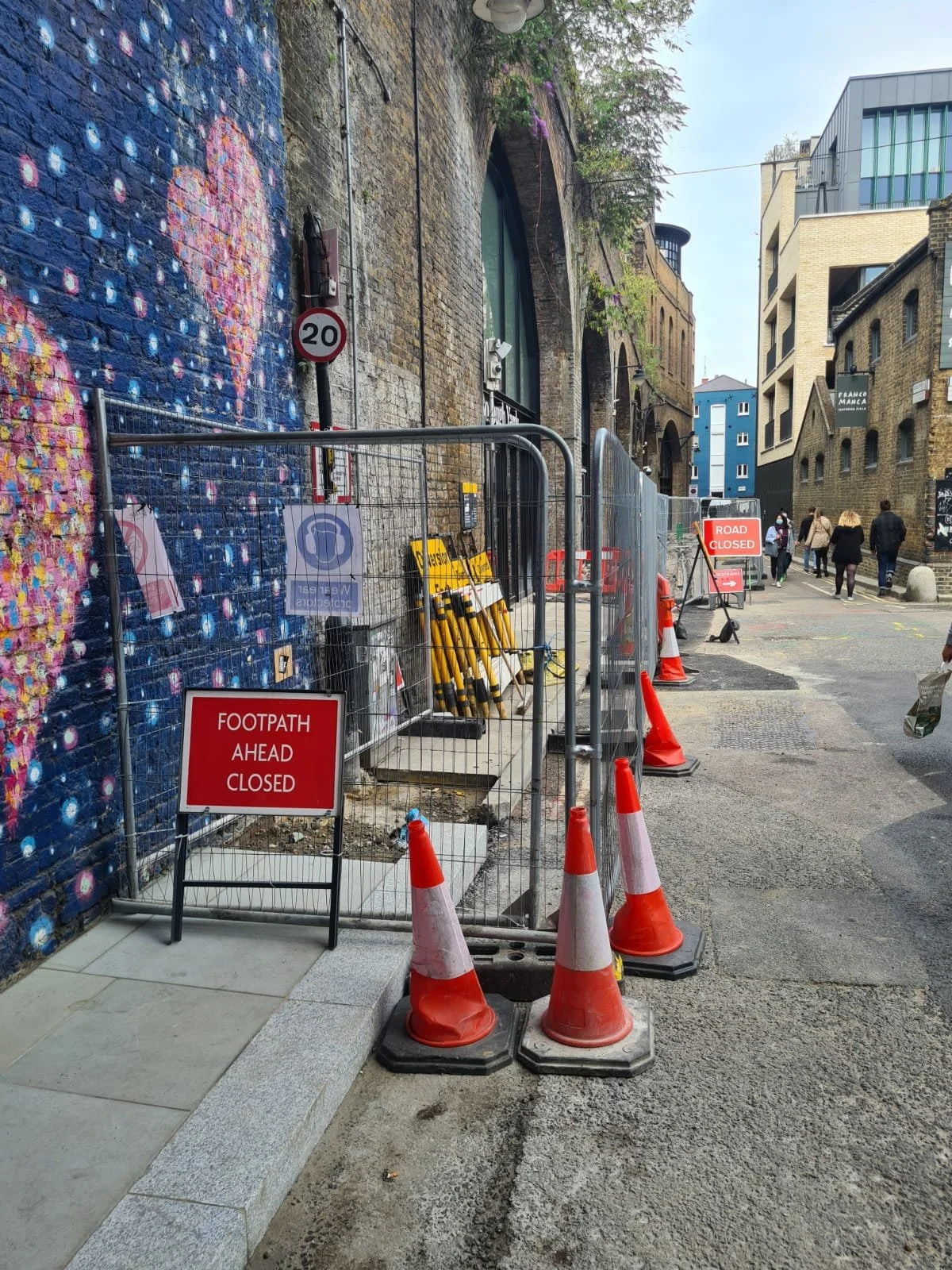 Street scene with construction barriers, orange cones, and signs indicating footpath and road closure, with pedestrians walking on the sidewalk and the background showing brick and modern buildings.