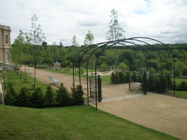 A gated garden with arched metal framework, surrounded by lush green grass and trees, with a view of a park in the background.