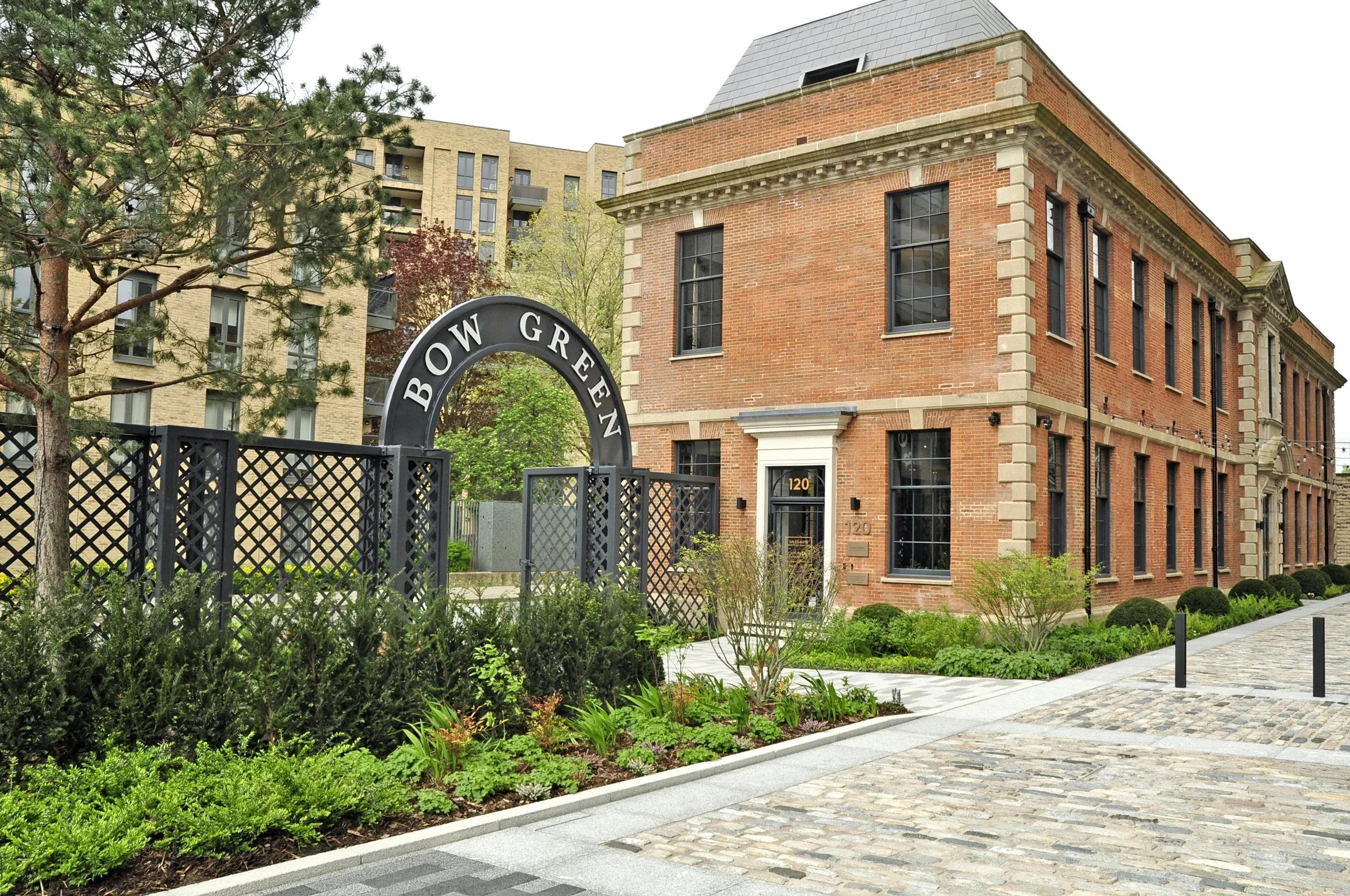 View of a modern red brick building with black-framed windows, next to a black lattice fence with a sign that reads 'BOW GREEN', on a cobblestone street with landscaped greenery and trees.