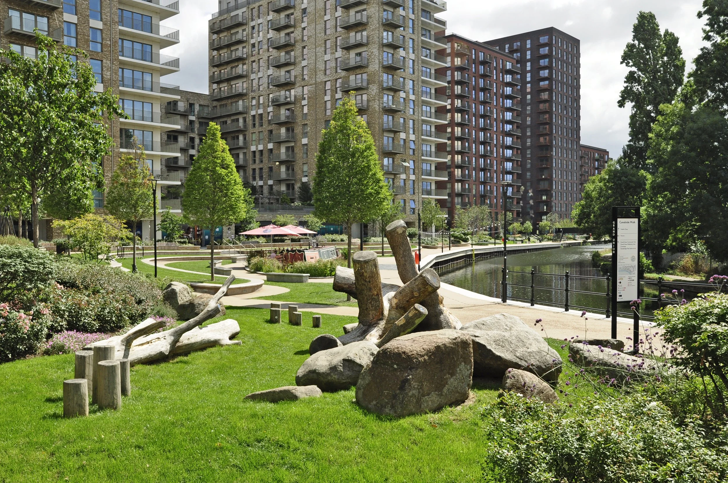A city park with green grass, trees, rocks, and wooden logs in the foreground. Behind, there are modern high-rise apartment buildings and a waterway with a railing on the right. A sign with park information is visible near the water.