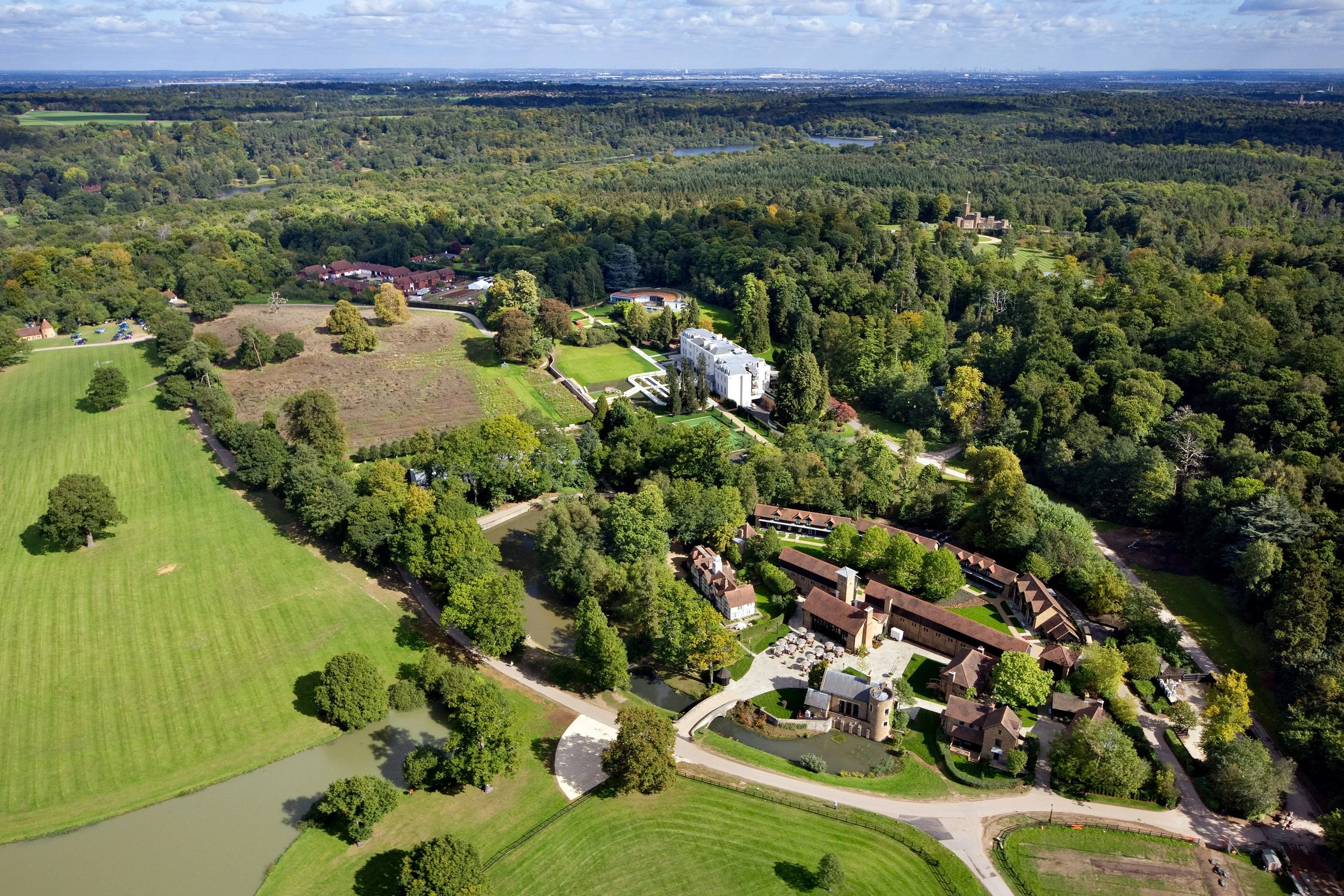 Aerial view of a large estate surrounded by trees, farmland, and a small pond, with a mix of modern and historic architecture, roads, and lush green landscape.