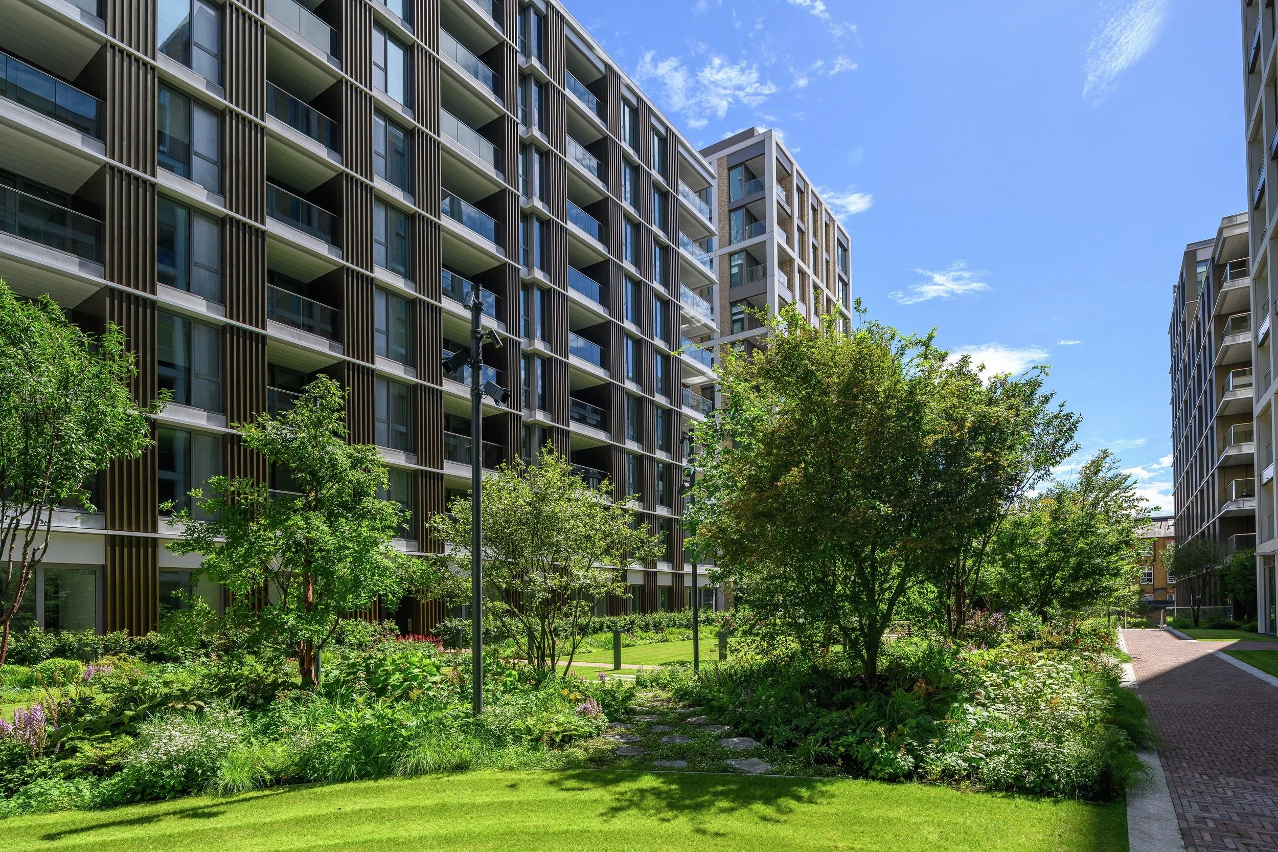 Modern residential apartment complex with landscaped garden, pathway, and trees under a blue sky.