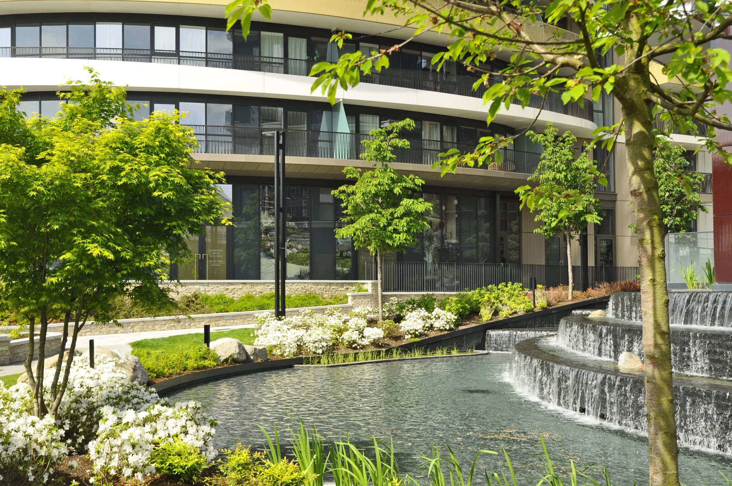 Modern apartment building with glass windows and balconies, surrounded by landscaped garden with several trees, flowers, a water fountain, and a small waterfall.