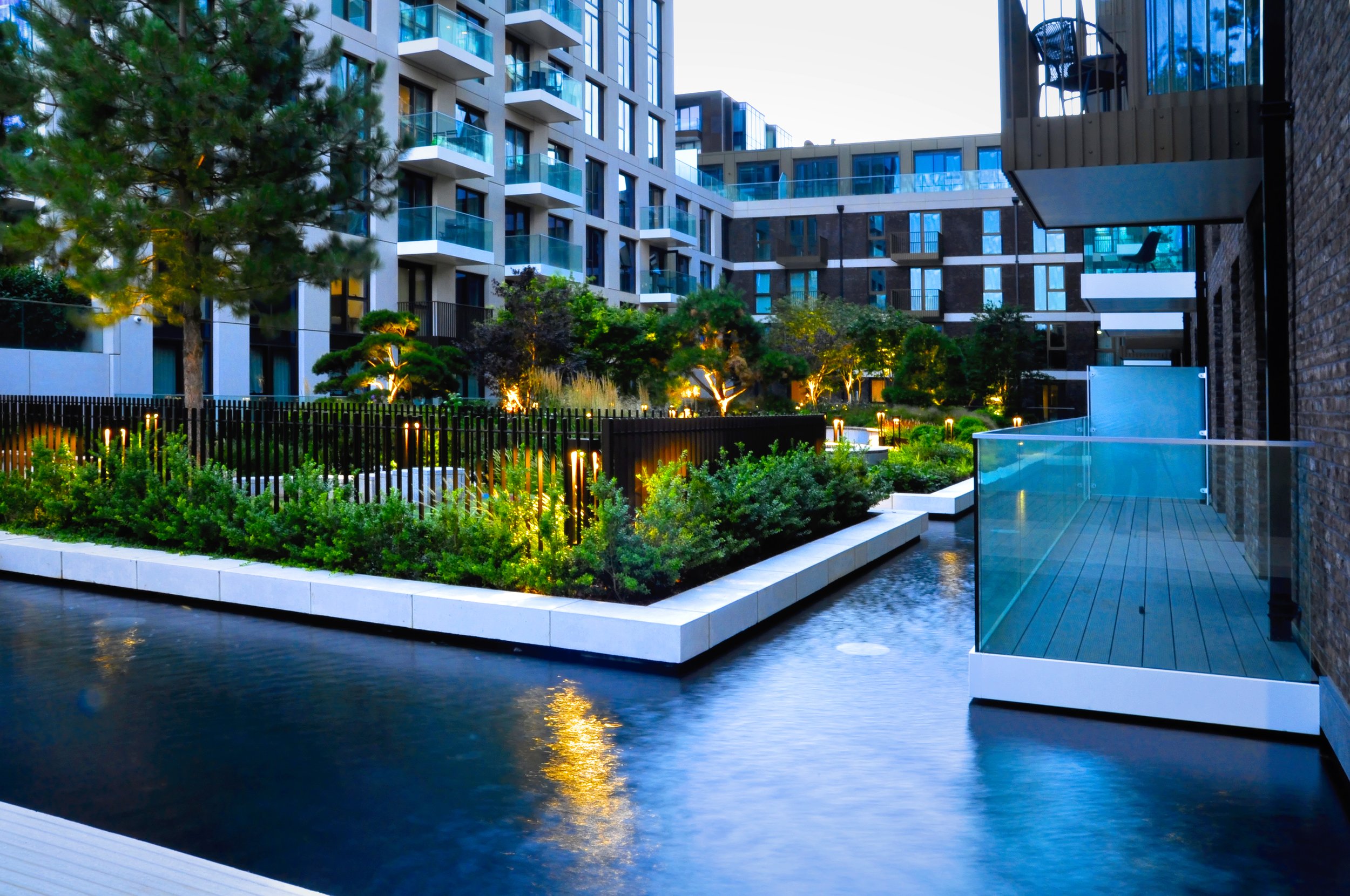 Modern residential apartment complex with a landscaped courtyard, water feature, and outdoor lighting at dusk.