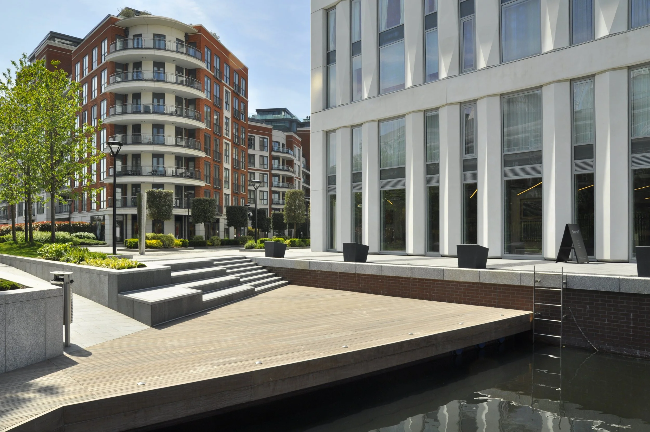 Modern urban waterfront area with a wooden dock, stairs, a small canal, trees, and contemporary multi-story buildings with glass windows and balconies.