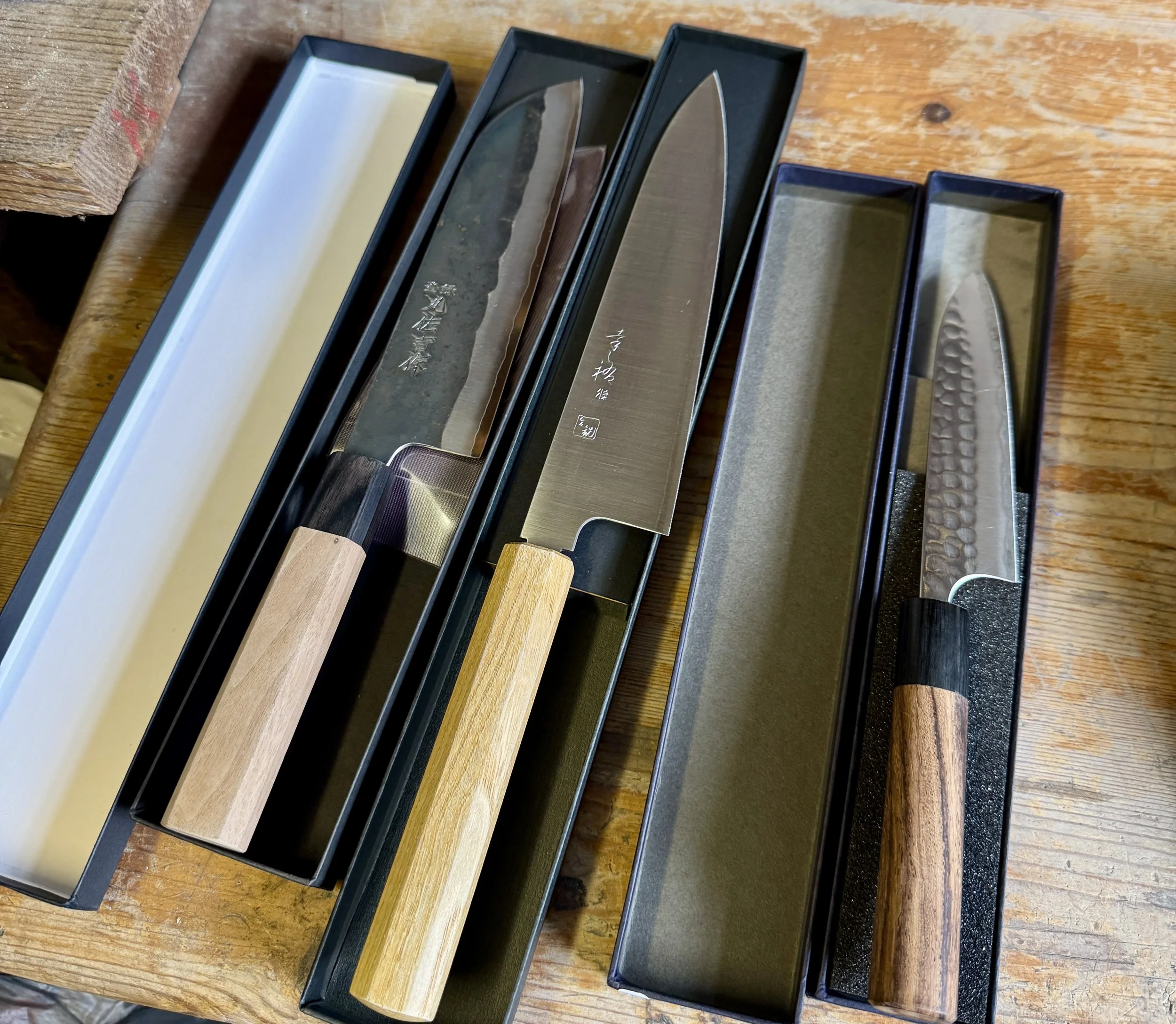 Collection of Japanese knives in black storage boxes on a wooden table.