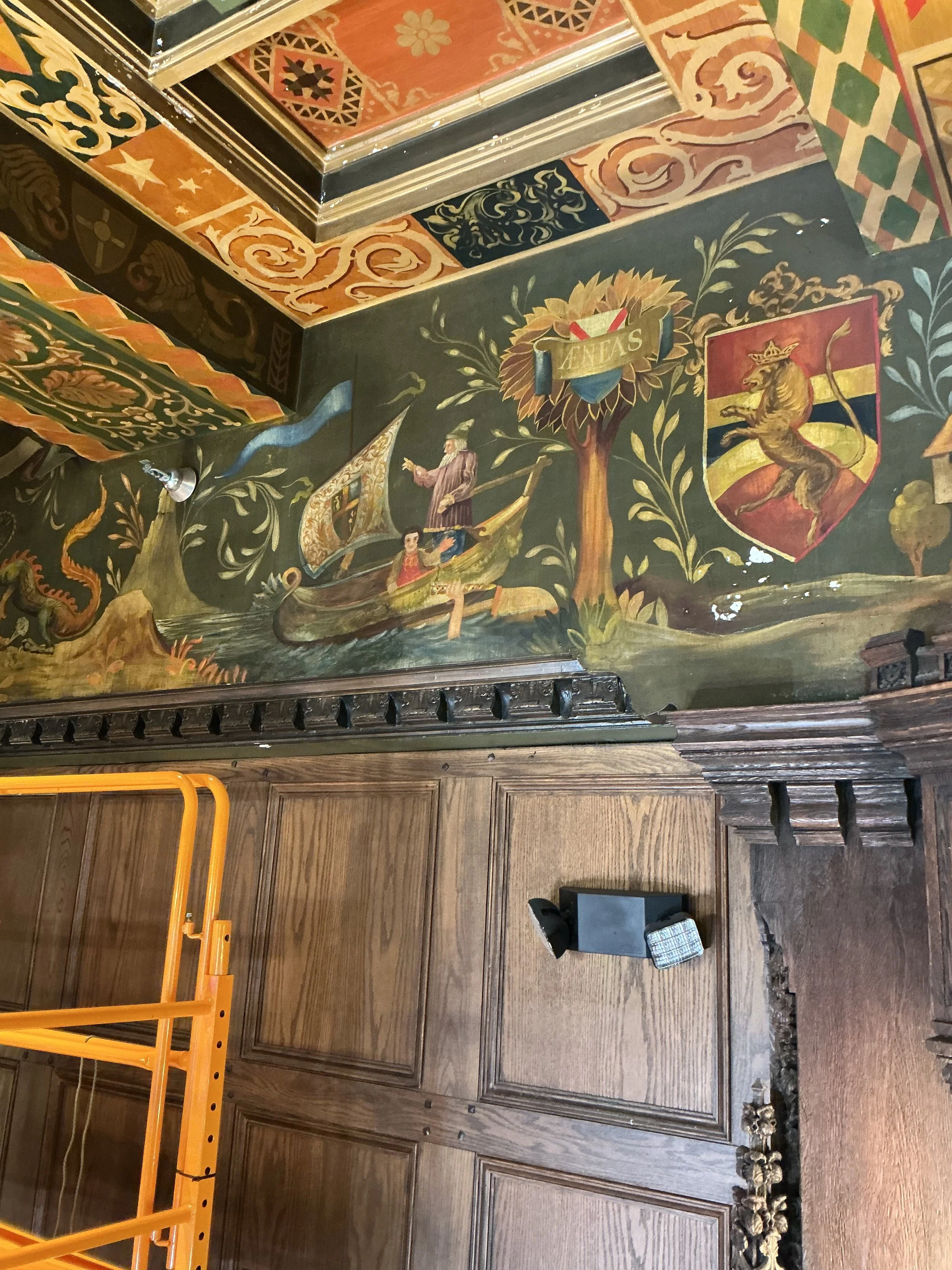 Decorative wall with painted shields and coat of arms, and a wooden panel below. A yellow metal frame with a ladder is on the left, a small black chair and a patterned rug on the right.