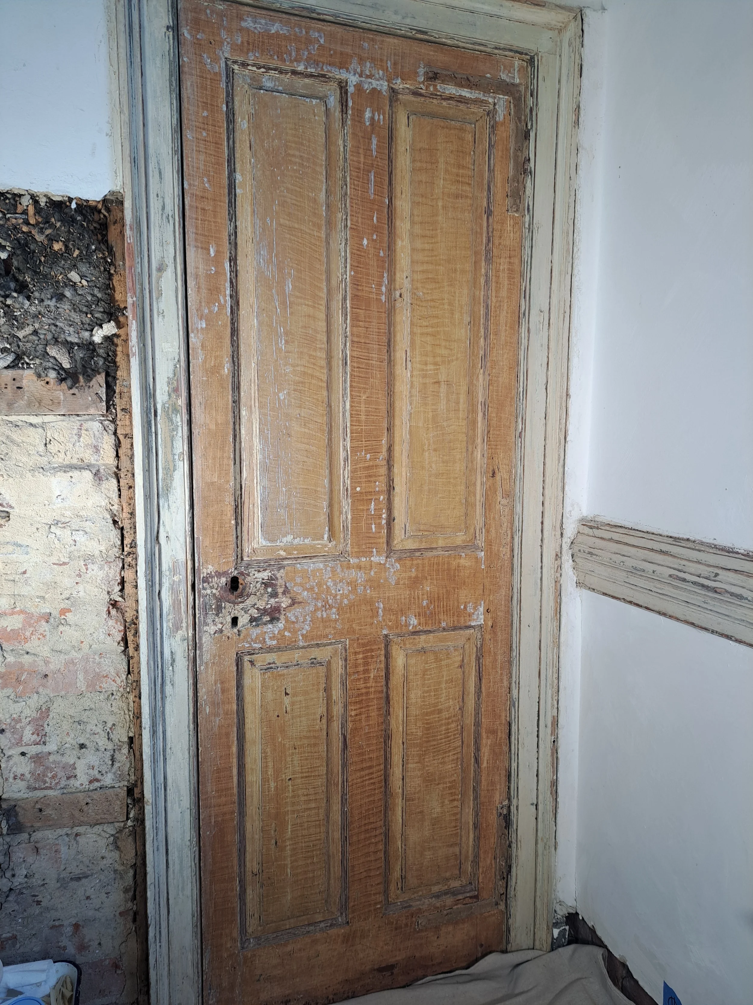 A wooden door with a distressed finish, revealing patches of worn paint and exposed wood, set within a white wall. The door has six rectangular panels and appears to be undergoing renovation.