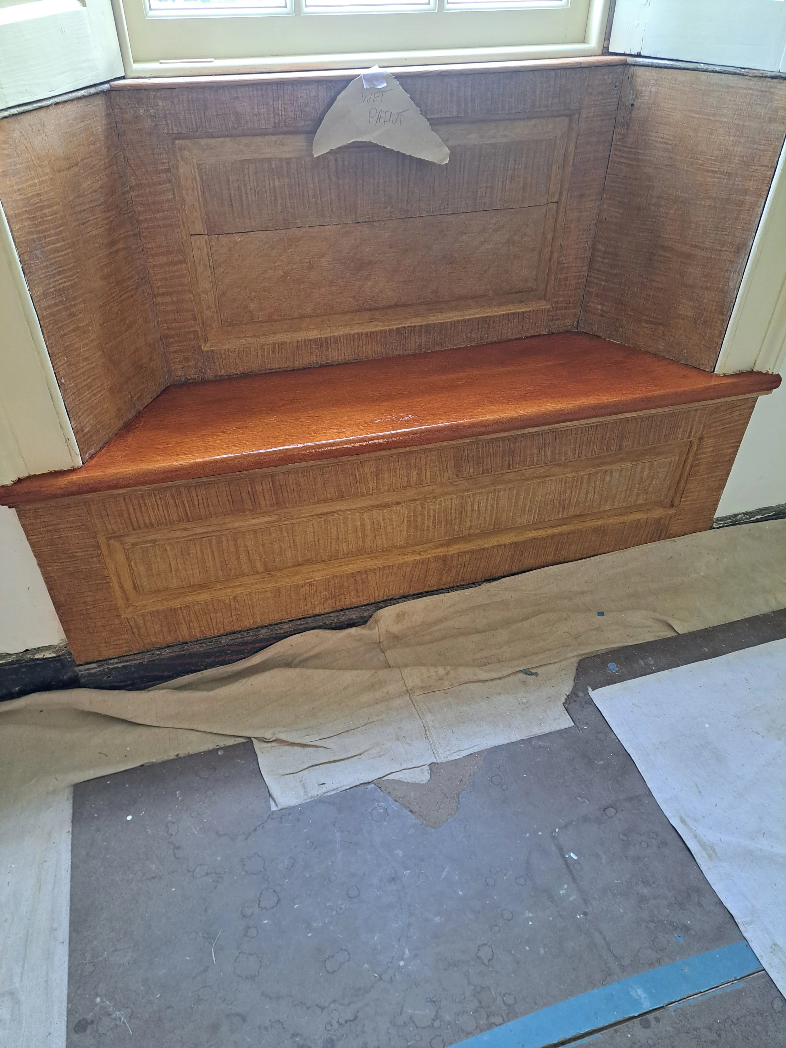 A wooden window seat with a panel at the back and a flat top, situated next to a window. A torn piece of paper with the handwritten note 'WET PAINT' is attached to the window.
