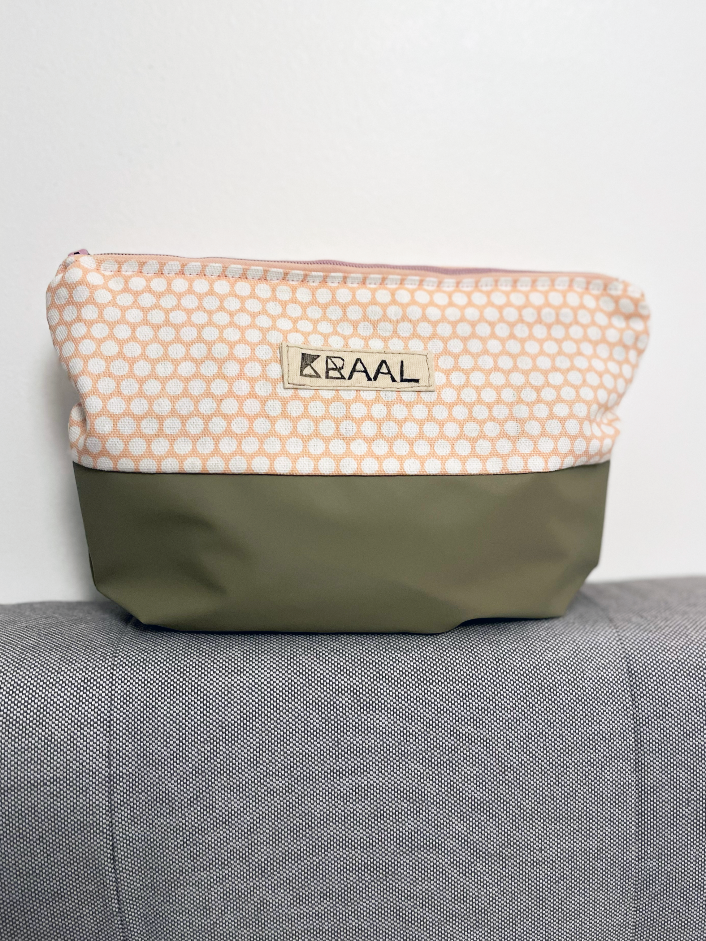 A pink and gray BAAL zippered pouch resting on a textured gray surface, with a white wall in the background.
