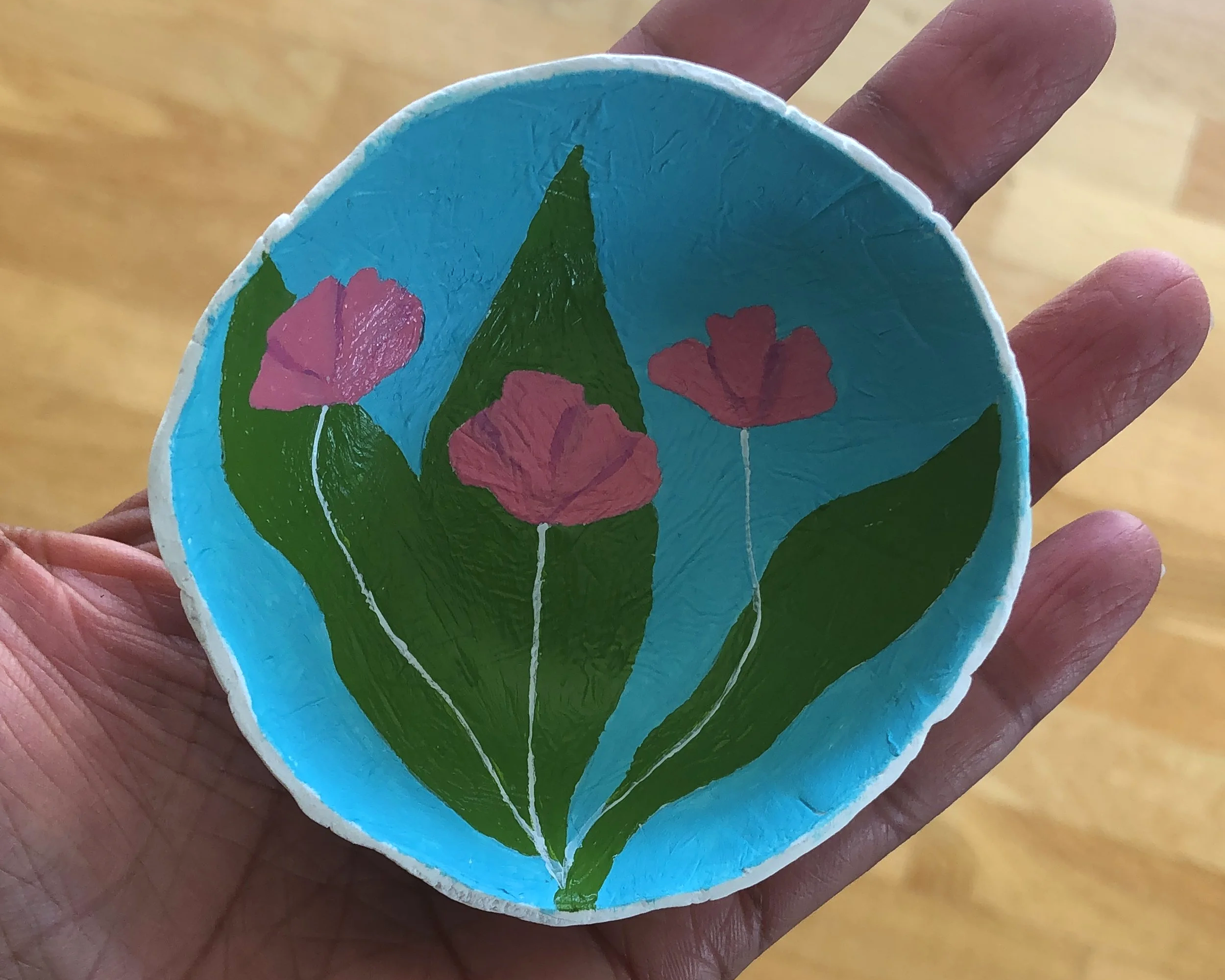Hand holding a round painted rock with three pink flowers and green leaves on a blue background.