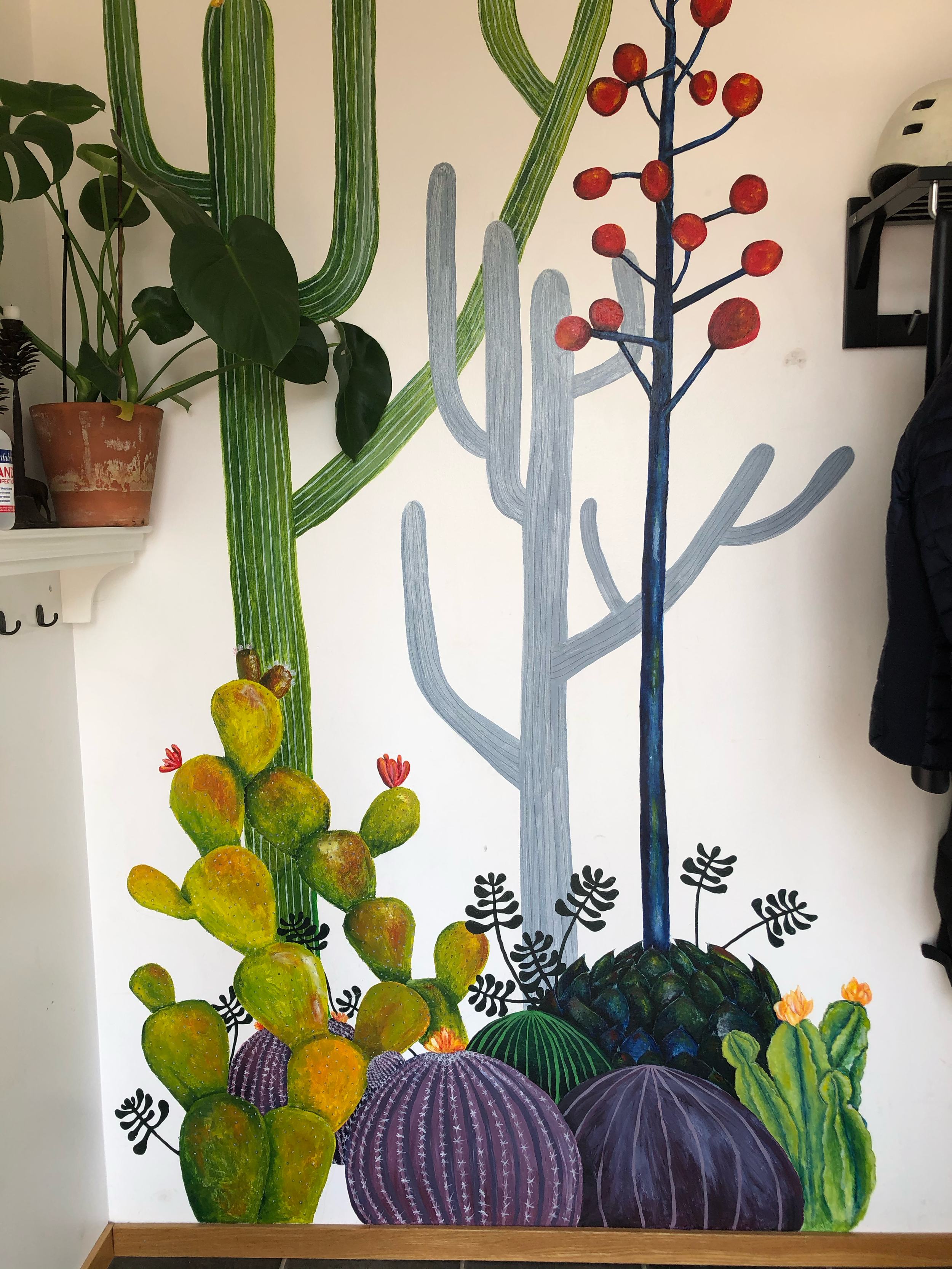 Colorful mural of various cacti and succulents painted on a white wall, featuring tall green and gray cacti, a blue and red-branched cactus with red fruit, and several other colorful succulents at the base.