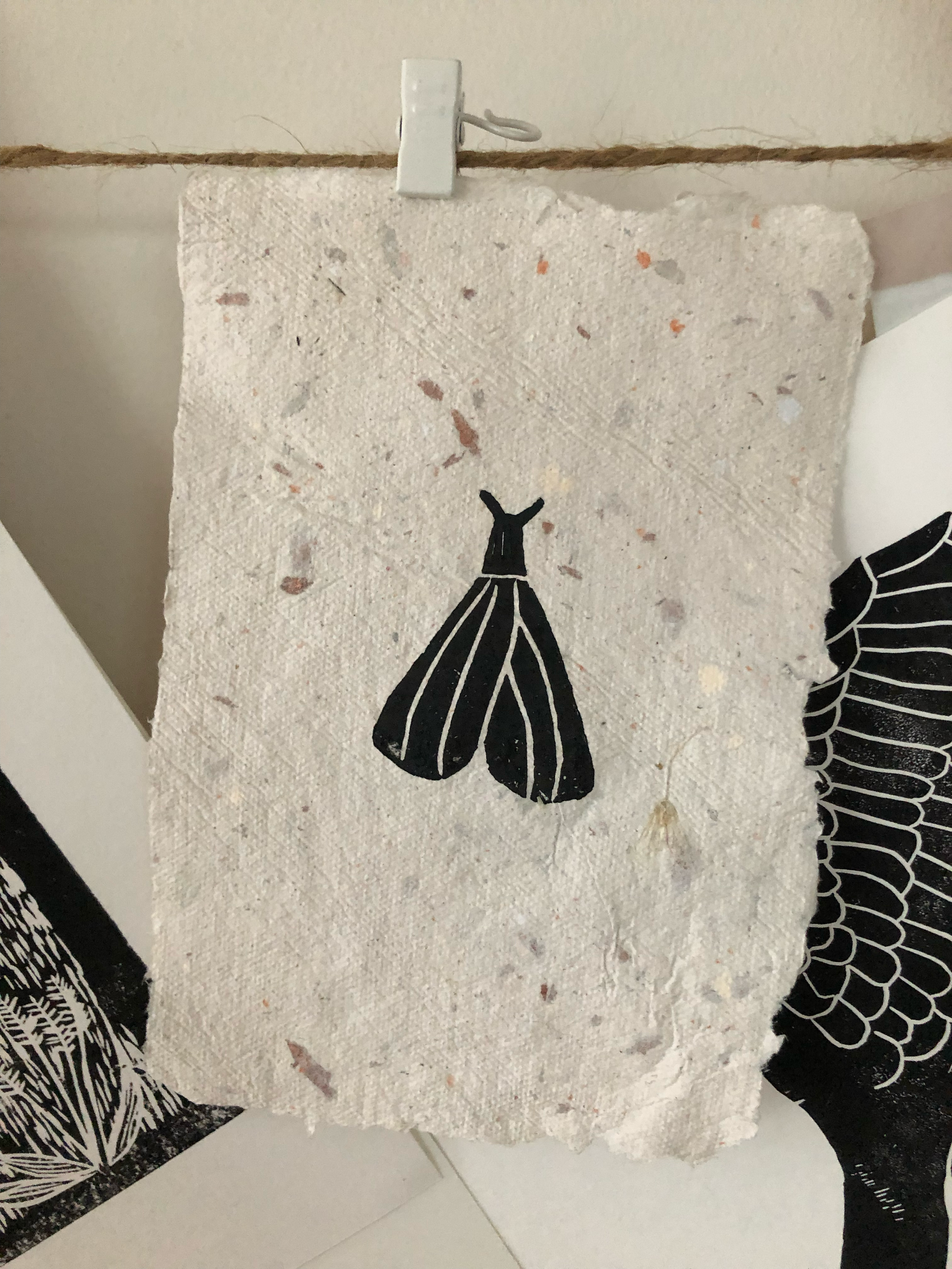 A piece of textured, cream-colored, handmade paper with a black silhouette of a moth printed on it, hanging from a string with a small white clip.