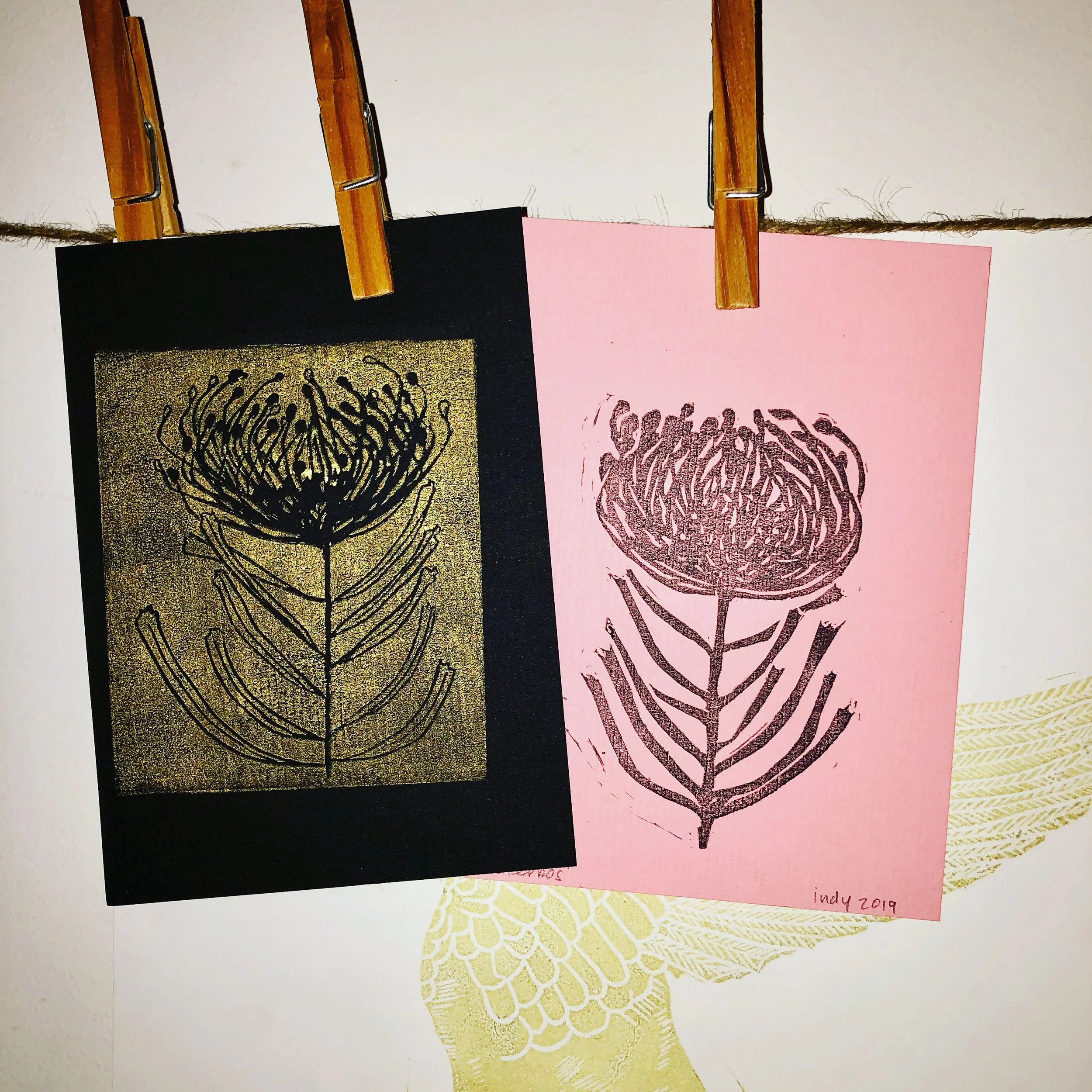 Two pieces of artwork featuring a stylized flower with a stem and leaves, hanging on a string with clothespins. One piece is on black paper with gold background, and the other is on pink paper with dark lines. Handwritten text on the pink paper reads