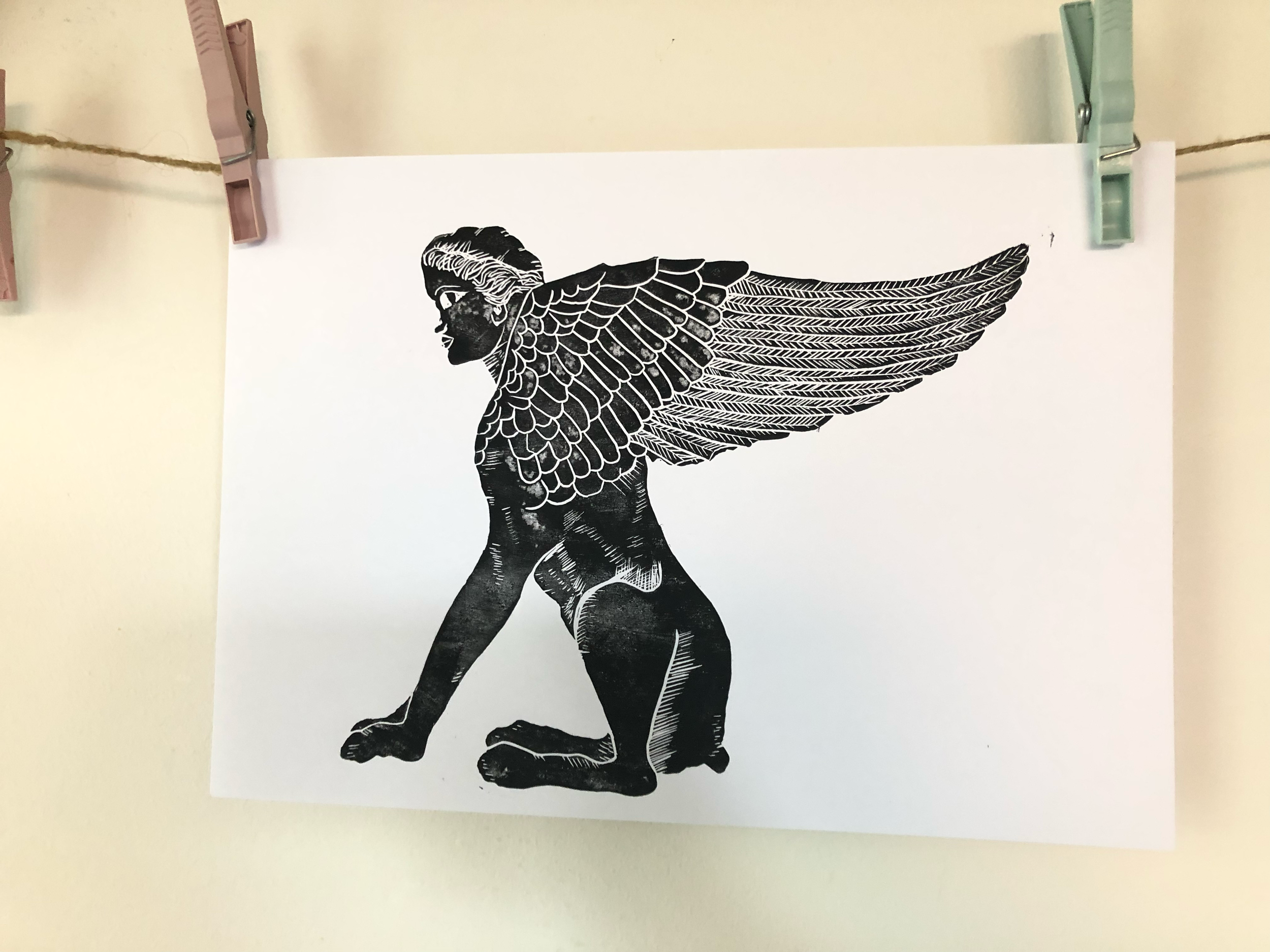 Black and white illustration of a mythological creature with the body of a lion, the head of a human, and large detailed bird wings, hanging on a wall with clothespins.