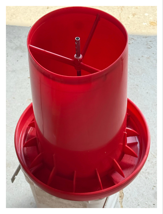 Red plastic bird feeder with a central tube and a circular perch around it, placed outdoors on a concrete surface.