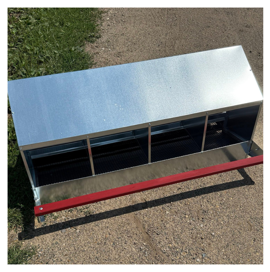 A metal outdoor chicken coop with four individual compartments on a gravel surface, with a grass field nearby.