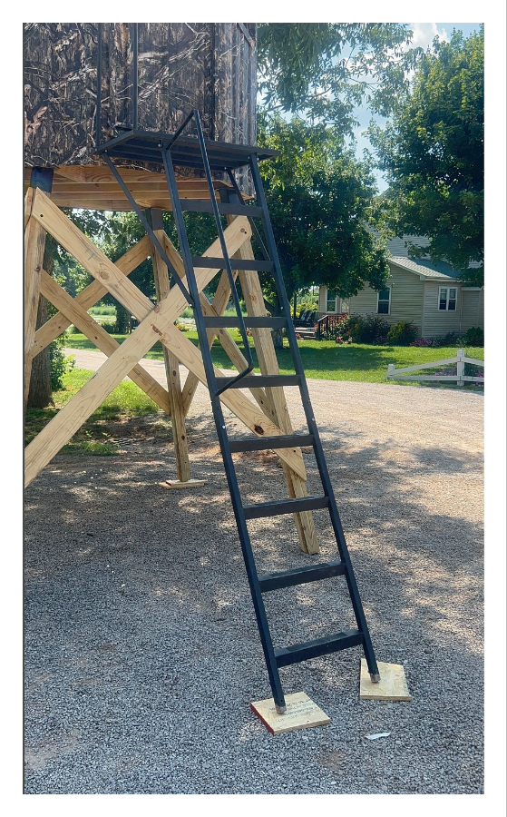 A black metal ladder leaning against a raised treehouse with camouflage siding, supported by wooden beams, and set on a gravel ground in a suburban backyard with trees and a house in the background.